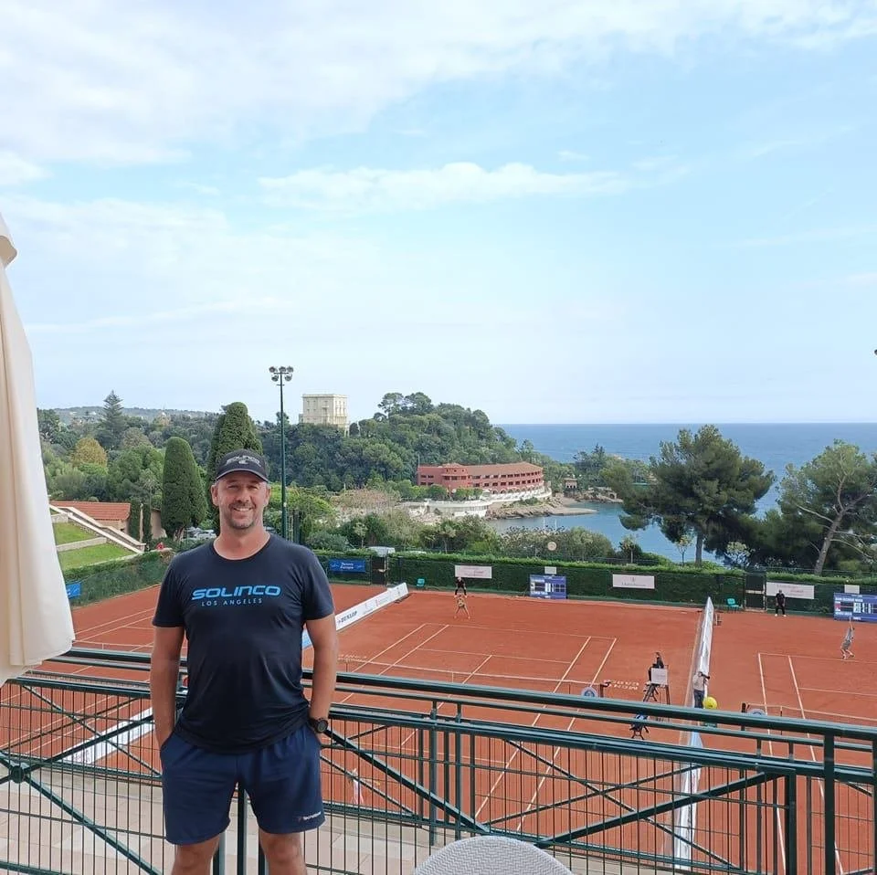 A man stands on a balcony overlooking a tennis court with the ocean and trees in the background.