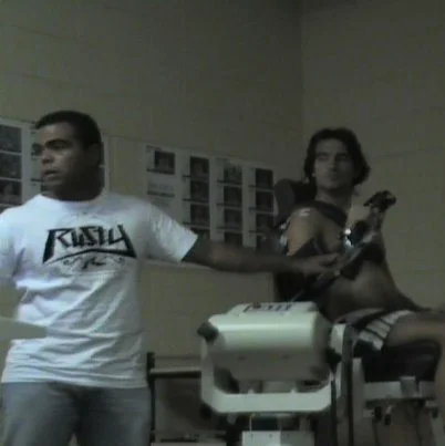 A man and a woman in a laboratory or medical setting. The man is holding an object connected to a device, and the woman is lying on a medical table holding a phone or small device.