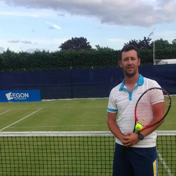 A man holding a tennis racket on a grass tennis court with a blue fence and advertising banner in the background.