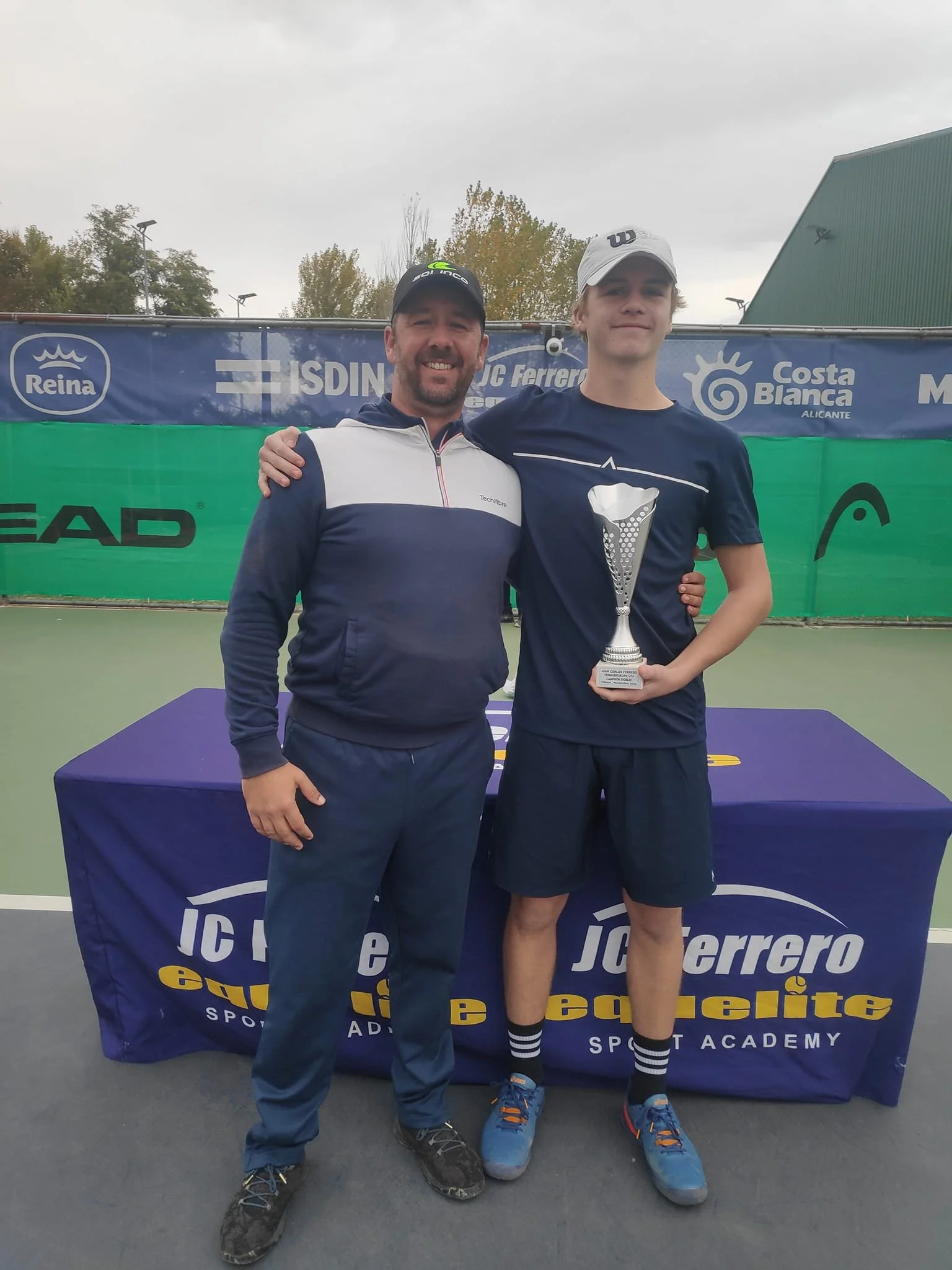 A man and a teenage boy standing on a tennis court, smiling at the camera. The teenager is holding a silver trophy. Behind them is a table with a blue cover that reads 'J.C. Ferrero' and 'Sport Academy'. In the background, there are banners with logos and a cloudy sky.