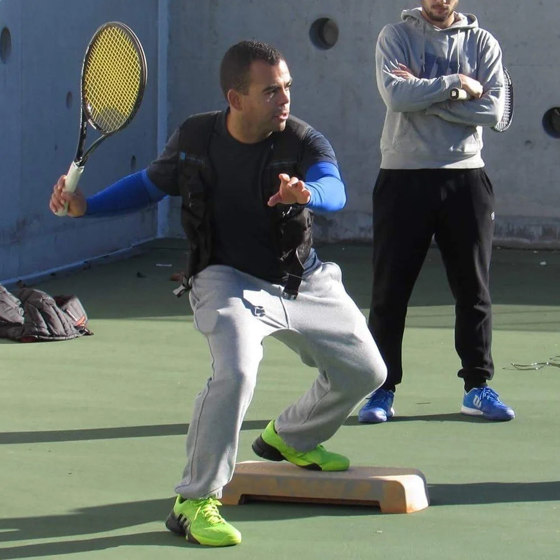 A man practicing tennis fitness training on a green tennis court with a balance board, holding a tennis racquet, while another man stands nearby observing.