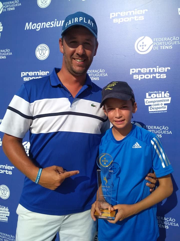 A man and young boy smiling at a tennis award ceremony, with the boy holding a trophy, against a blue backdrop featuring various sponsor logos and the Portuguese Tennis Federation logo.