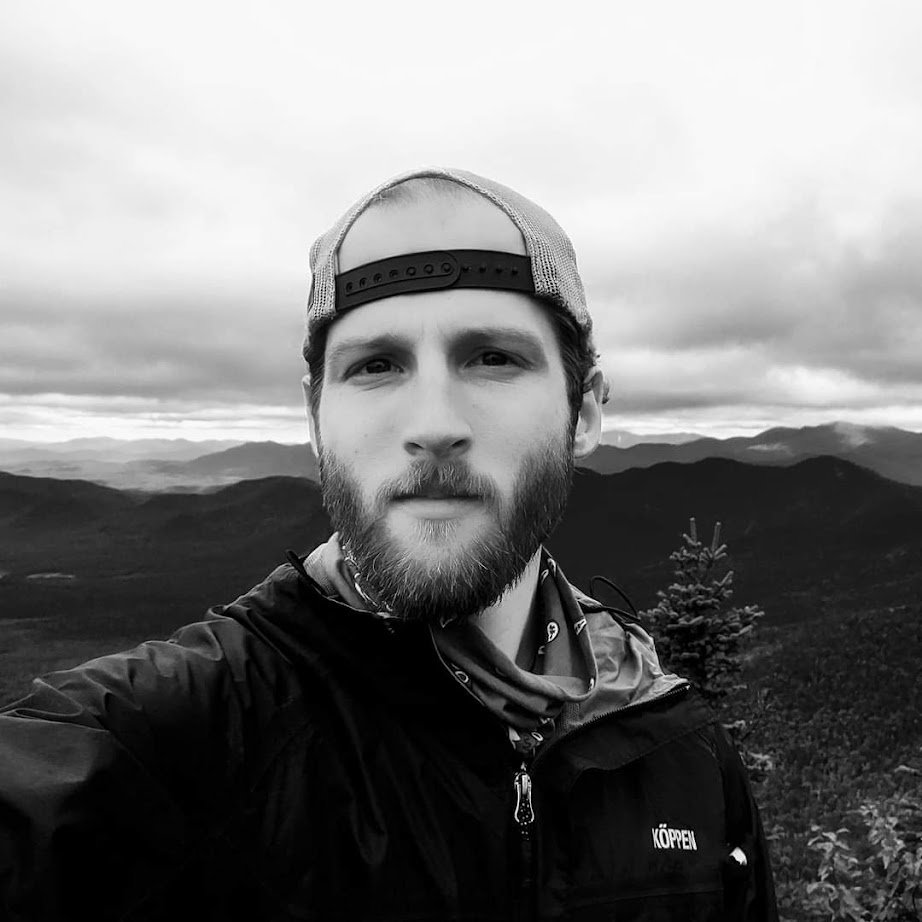 A man on a mountain during daytime with other mountains and a cloudy sky in the background.