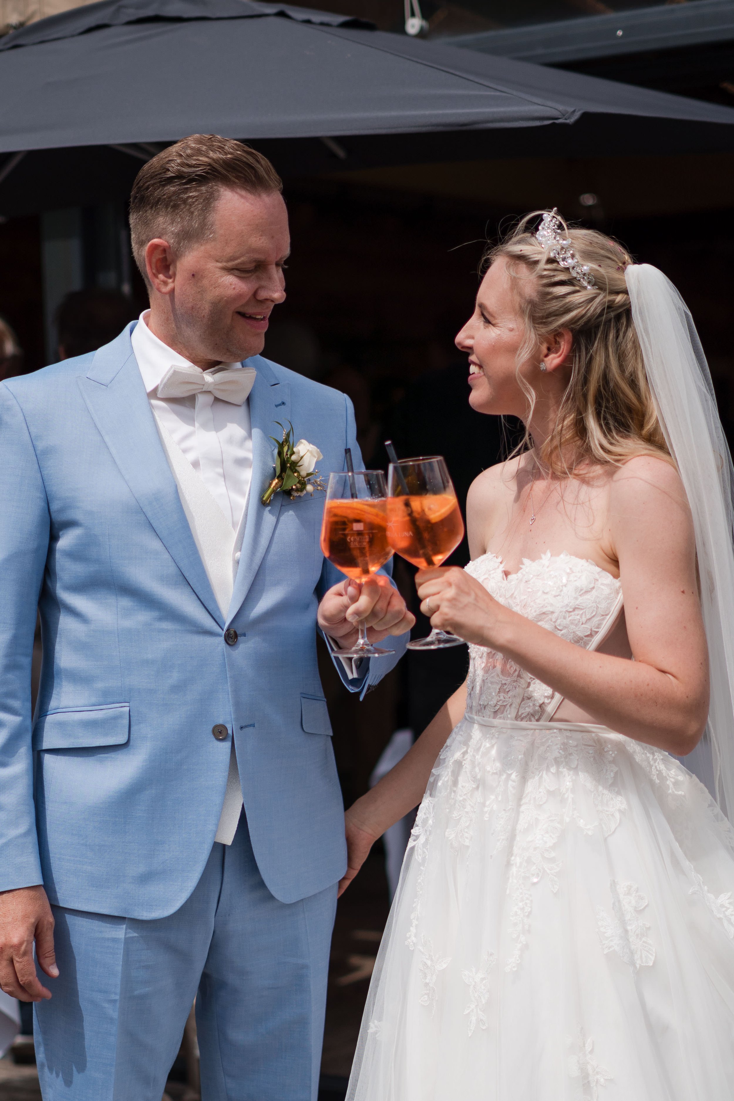 Ein frisch verheiratetes Paar beim Anstoßen mit einem Glas Aperol Spritz, die Braut trägt ein weißes Hochzeitskleid und trägt eine Tiara, der Bräutigam trägt einen hellblauen Anzug mit Fliege.