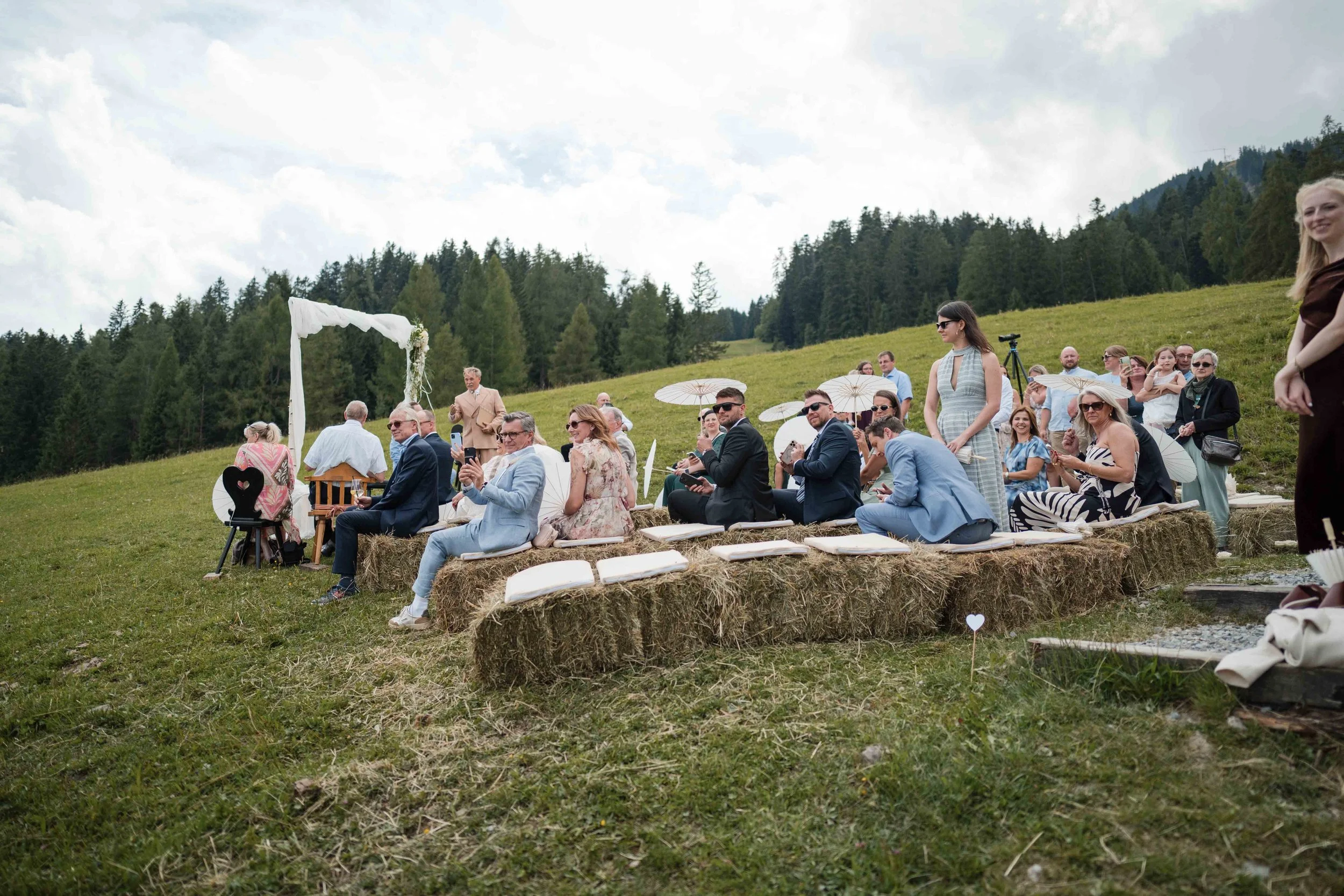 Menschen bei einer Hochzeitszeremonie im Freien, sitzend auf Strohballen, mit Dekorationen und Blick auf eine grüne Hügel- und Waldlandschaft.