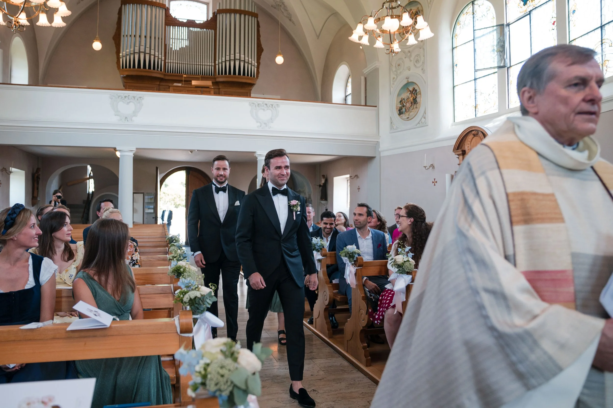 Zwei Männer in schwarzen Anzügen mit Fliegen, eines davon trägt eine rosafarbene Blume, gehen in einer Kirche an der Hochzeitsszene vorbei, umgeben von Gästen, die auf den Bänken sitzen, mit Dekoration und einem Orgel im Hintergrund.