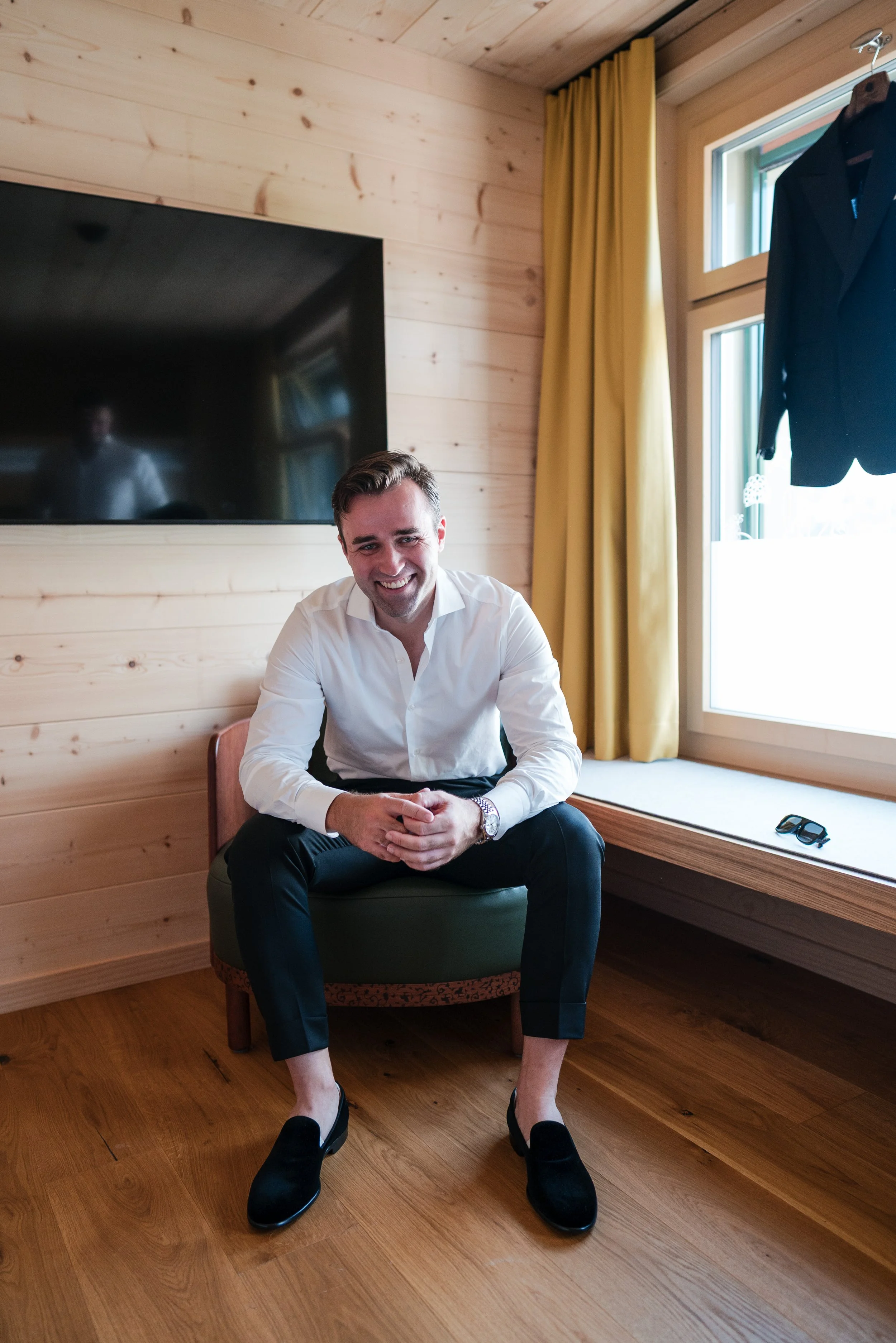 Ein lächelnder Mann in einem weißen Hemd und schwarzen Hosen sitzt in einem Holzhaus auf einem Stuhl, neben einem großen Fenster mit gelben Vorhängen, auf dessen Fensterbank eine dunkle Jacke und eine Sonnenbrille liegen.