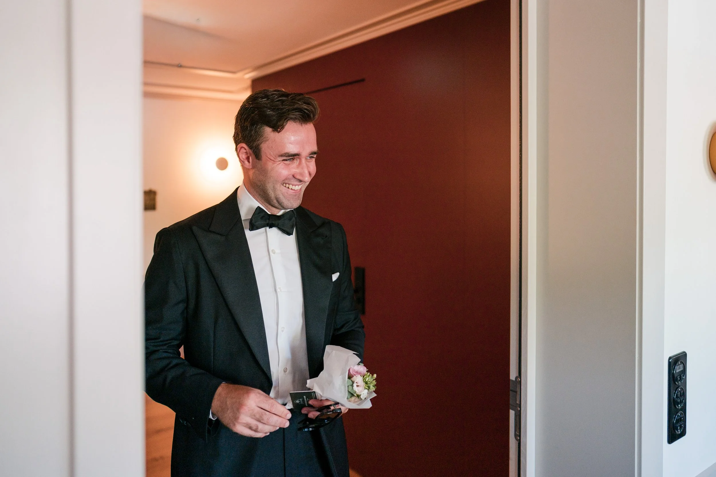 Ein Mann in Smoking mit Fliegenabzeichen hält einen kleinen Blumenstrauß und Schlüssel, steht vor einem Fahrstuhl in einem modernen Haus.
