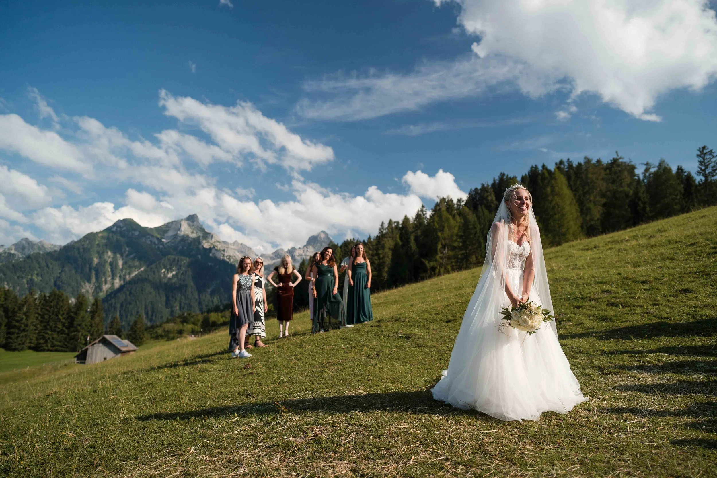 Braut im weißen Kleid mit Blumenstrauß, lachend auf einer Wiese, im Hintergrund eine Gruppe von Frauen, Berge und Himmel.