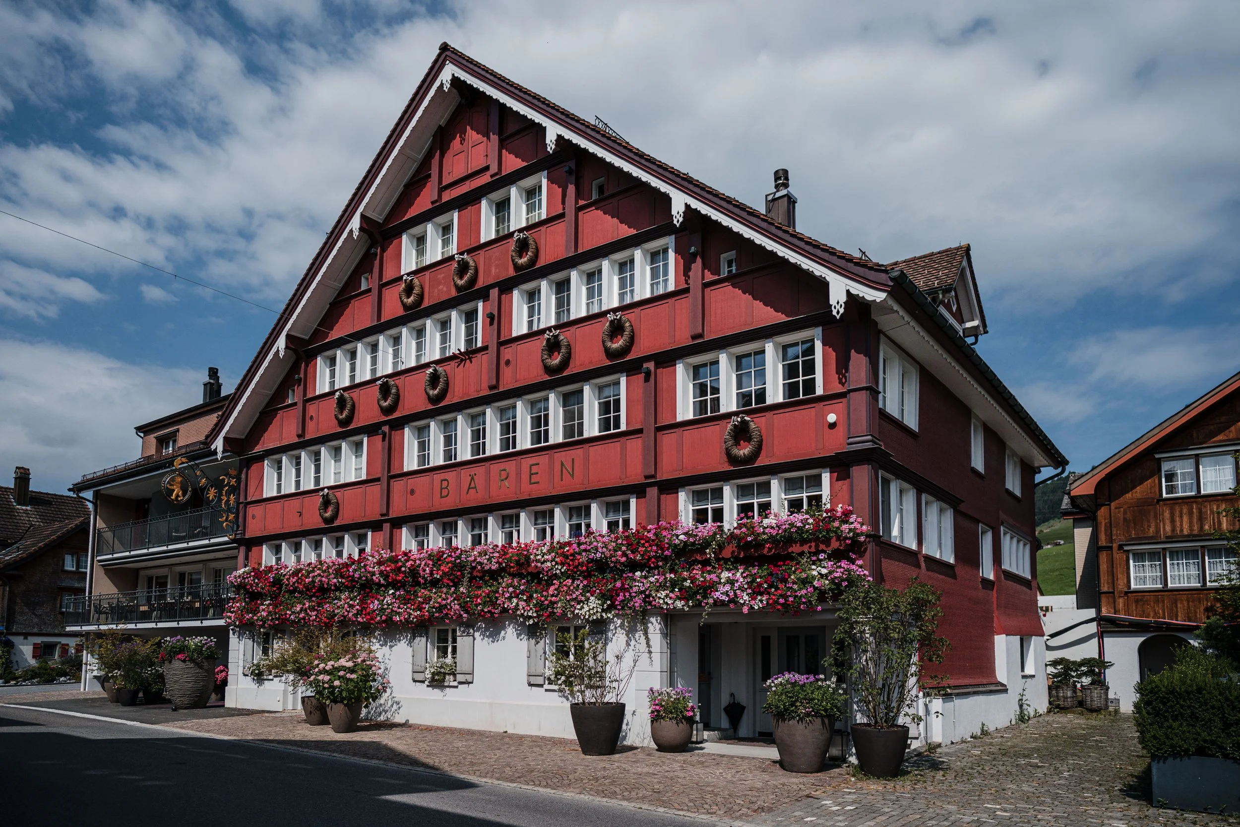 Ein mehrstöckiges, rotes Holzgebäude mit Blumen und Kränzen, in einer kleinen Stadt, mit dem Schriftzug "Bären" auf der Fassade.