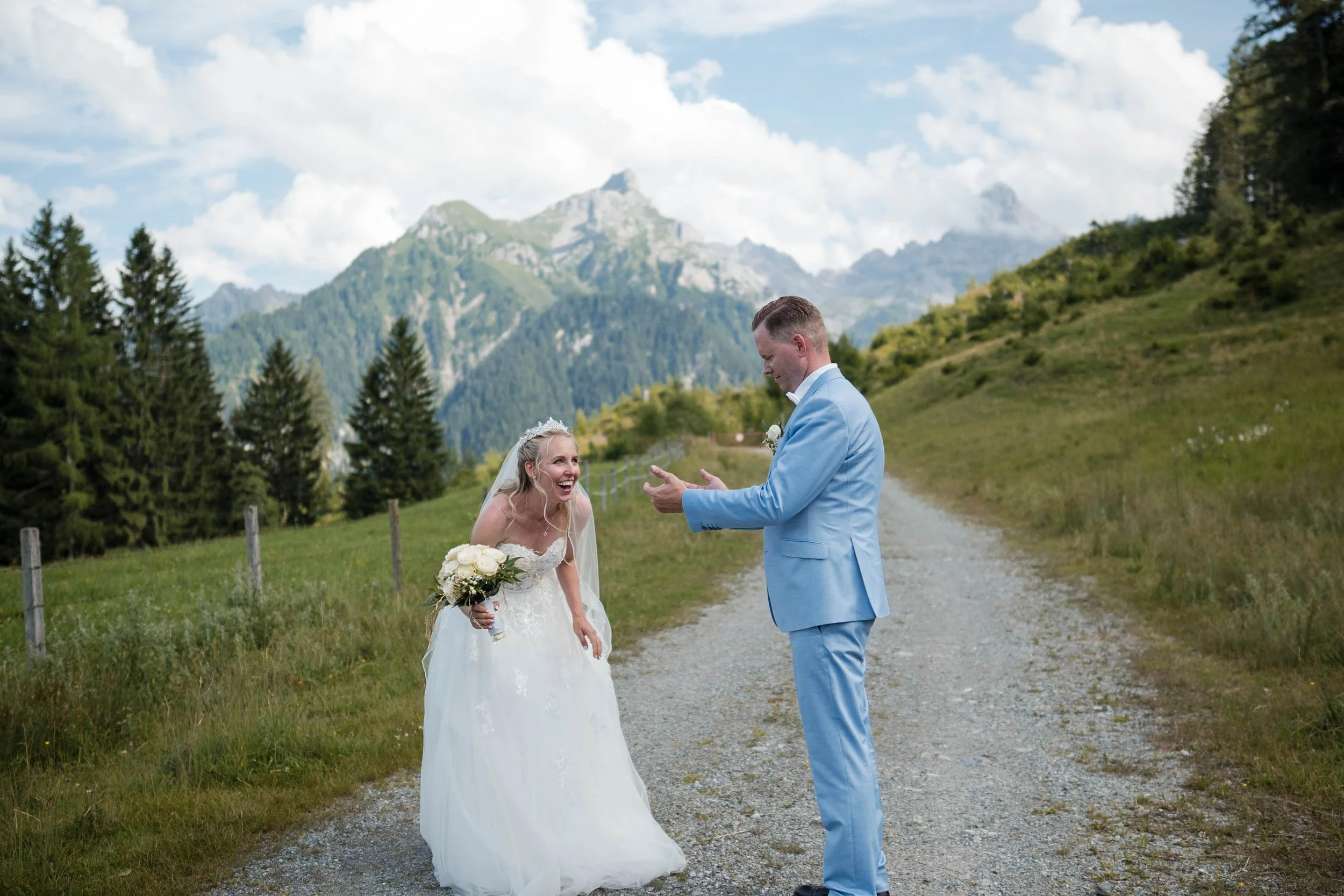 Ein Brautpaar steht auf einer Schotterstraße in einer malerischen Berglandschaft. Die Braut lacht, hält einen Blumenstrauß, während der Bräutigam in einem hellblauen Anzug vor ihr steht.