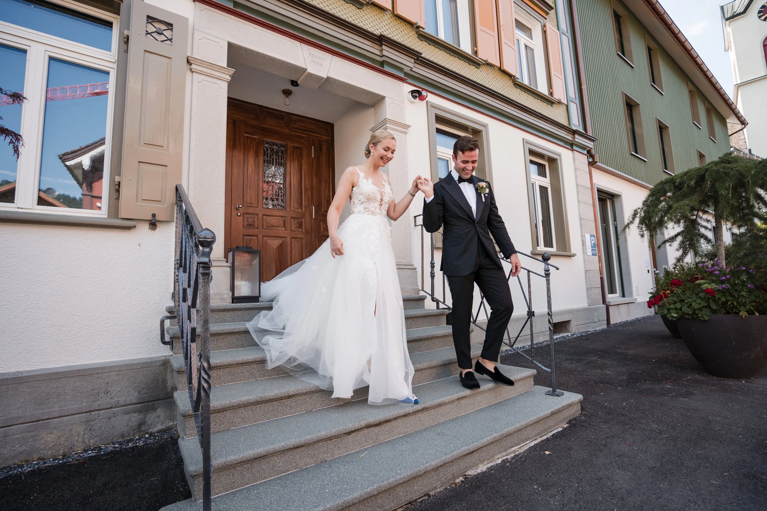 Braut und Bräutigam steigen Hand in Hand die Stufen eines Hauses hinunter, beim Hochzeitstag, vor einem Haus mit bunten Fassaden und begrünten Balkonen.