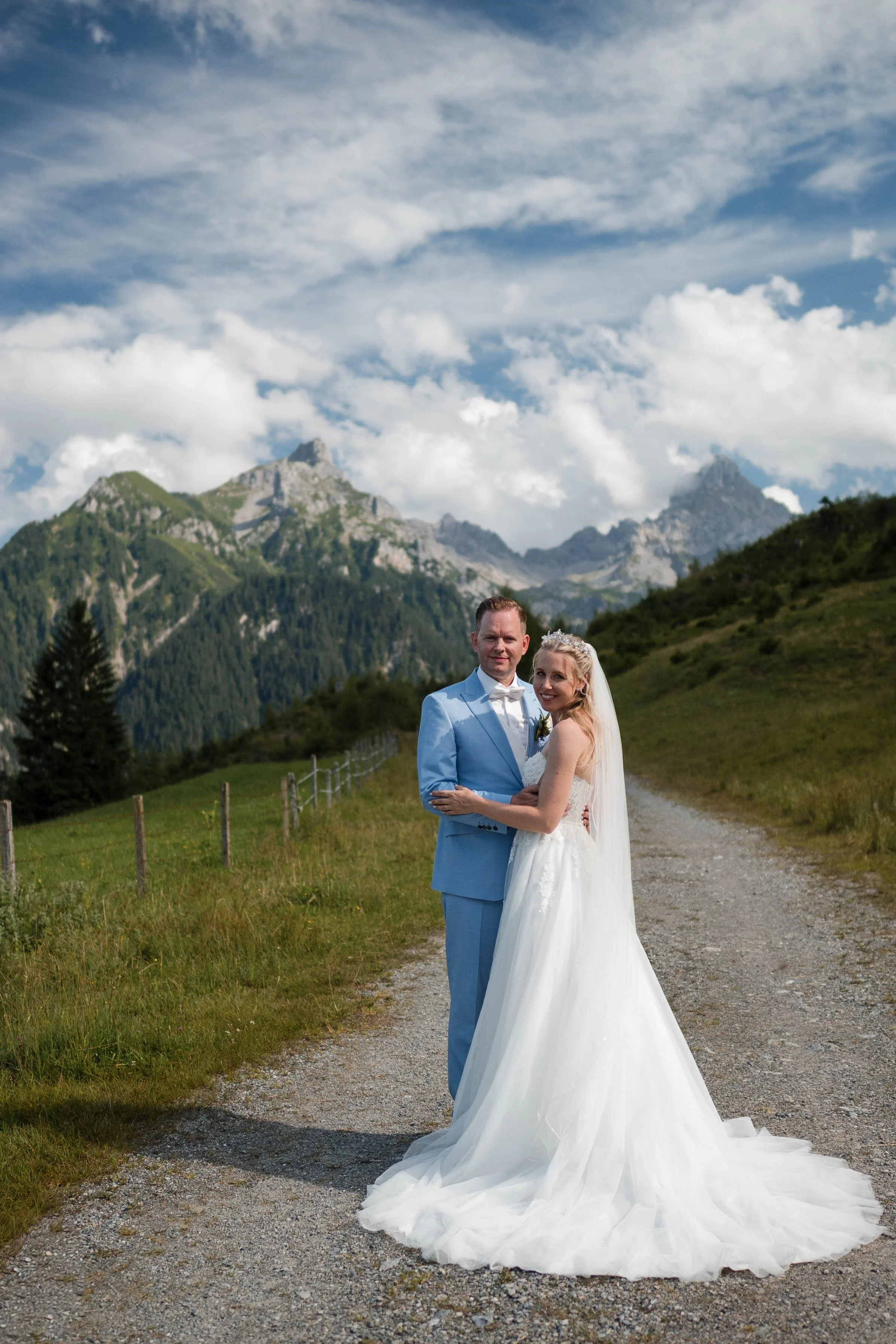 Ein Brautpaar auf einem Schotterweg in den Bergen, umgeben von grünen Wiesen und hohen Bergen bei Sonnenlicht, mit einem blauen Himmel und Wolken im Hintergrund.