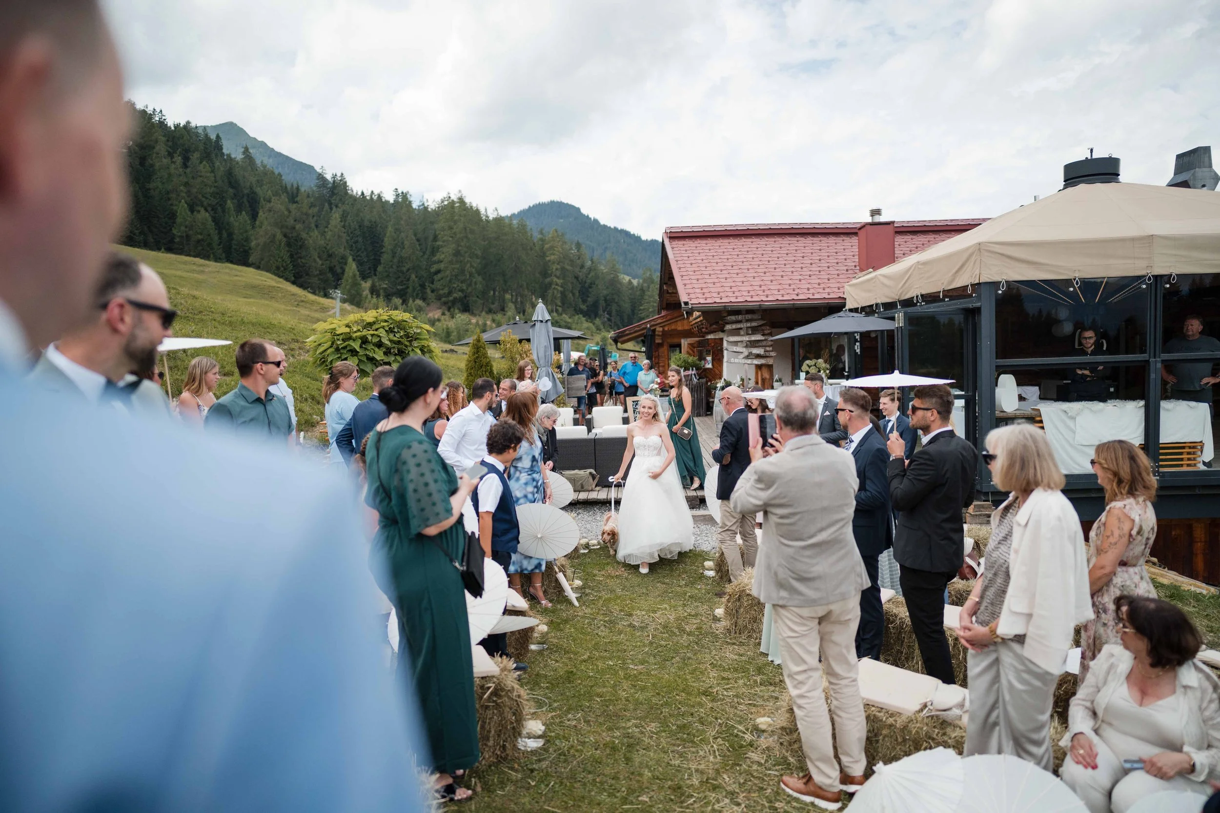 Hochzeit im Freien mit vielen Gästen, einer Braut in Weiß, umgeben von Bergen und Grünland, bei gutem Wetter