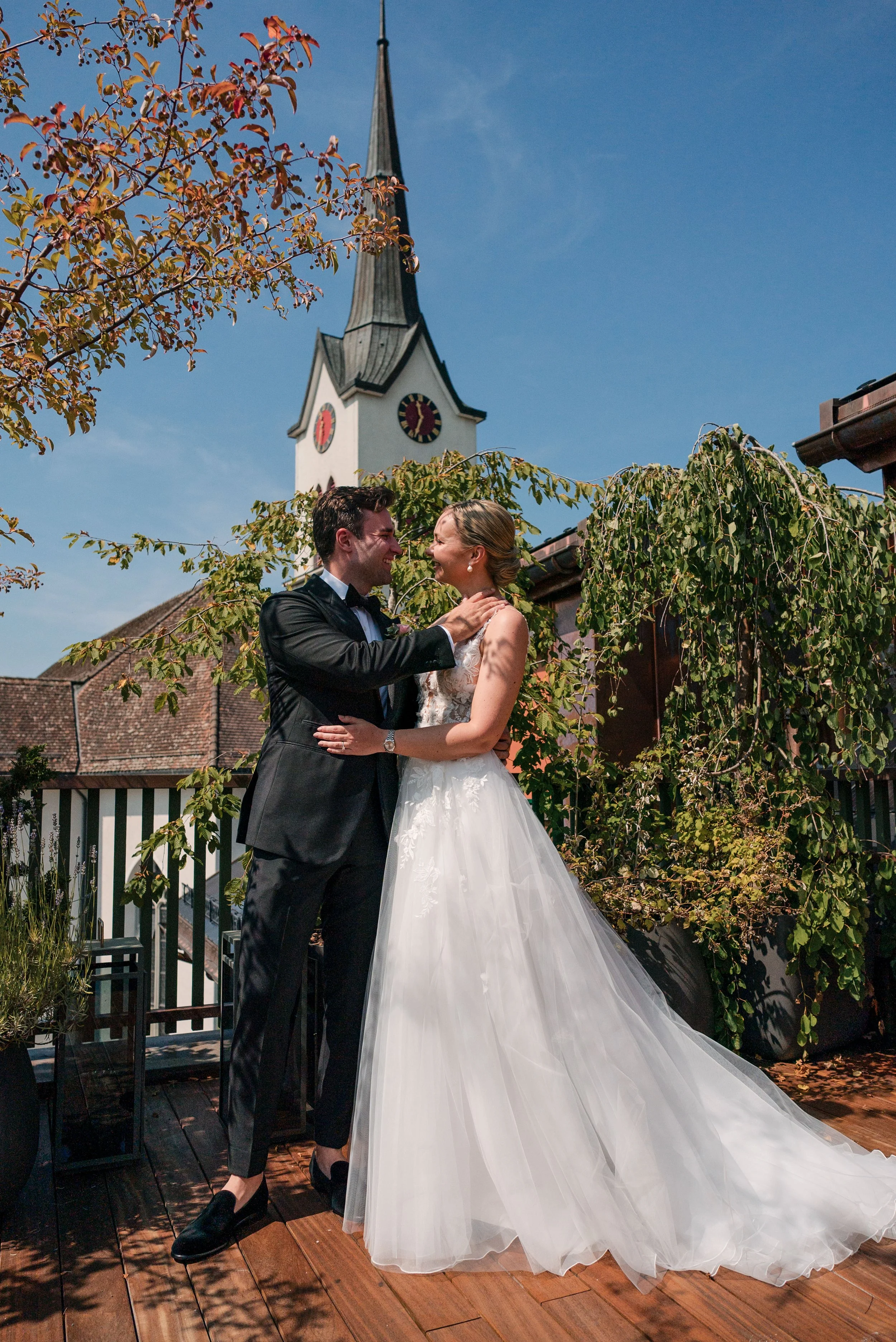 Ein Paar, das an einem sonnigen Tag auf einer Terrasse in der Nähe einer Kirche in einem Hochzeitskleid und einem Anzug posiert, umarmt sich und schaut sich lächelnd an.