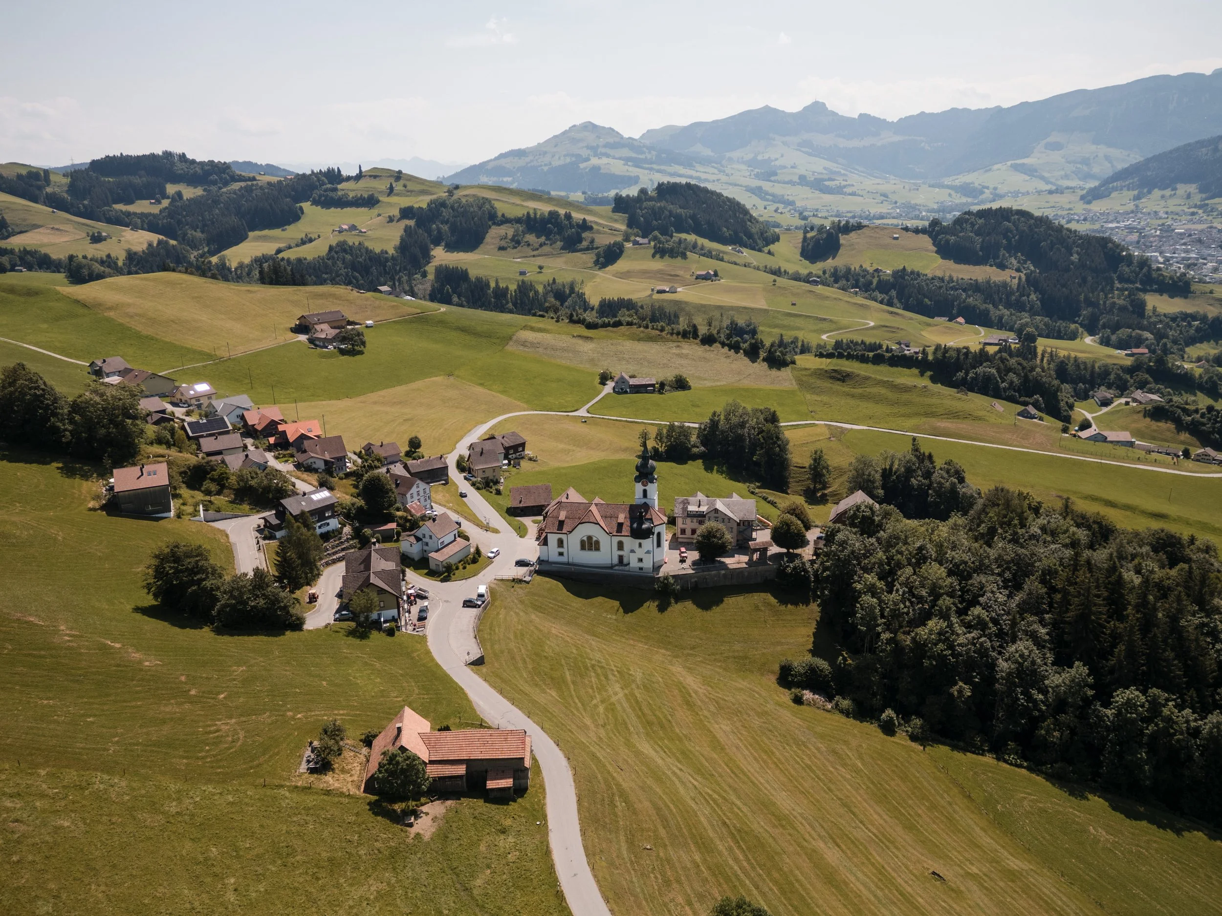 Luftaufnahme eines kleinen Dorfes inmitten grüner Hügel und Berge, mit einer Kirche im Zentrum.
