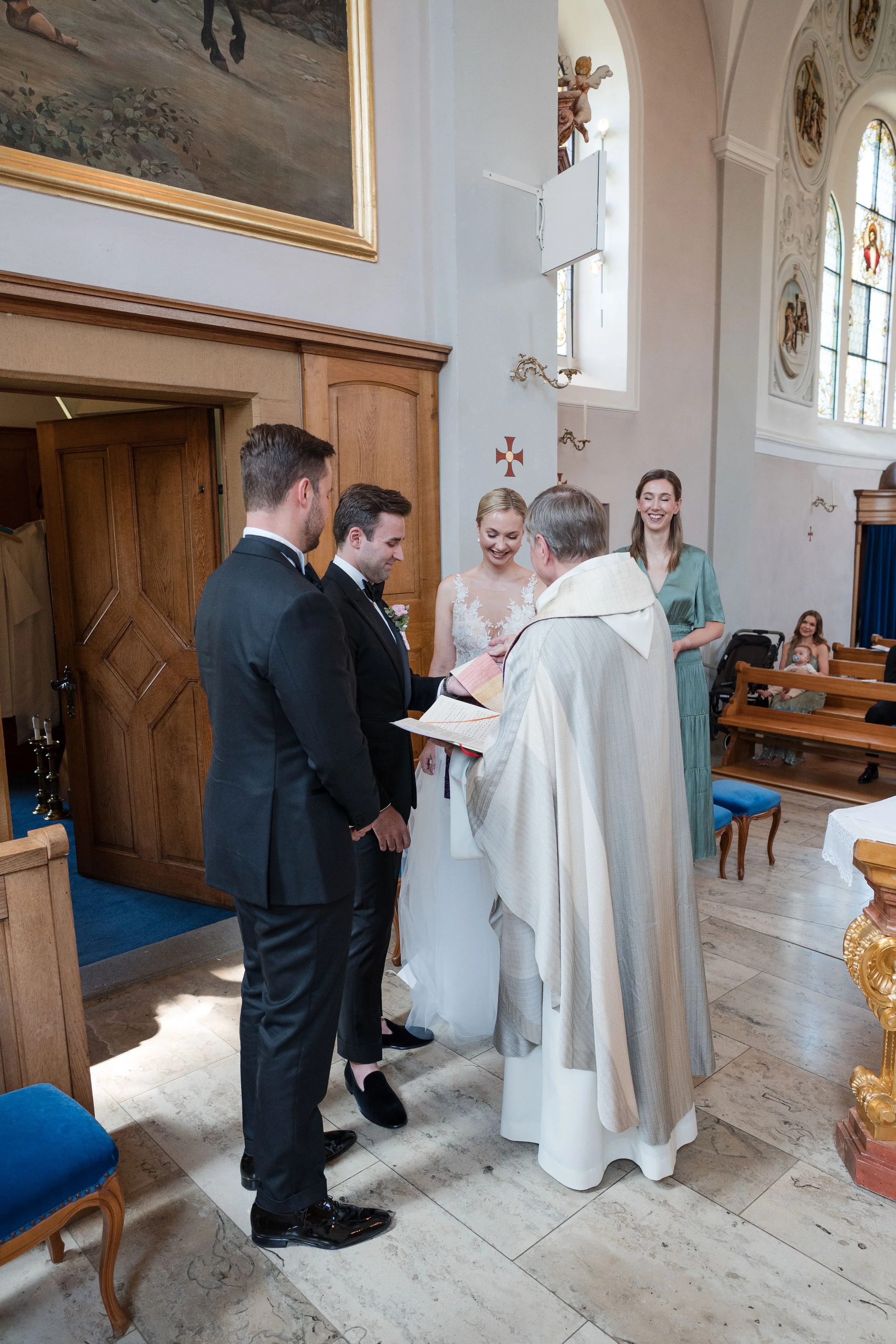 Hochzeit in einer Kirche, der Priester segnet das Brautpaar während der Zeremonie, umgeben von Gästen, mit Blick auf die Kirchenfenster und Wanddekorationen.