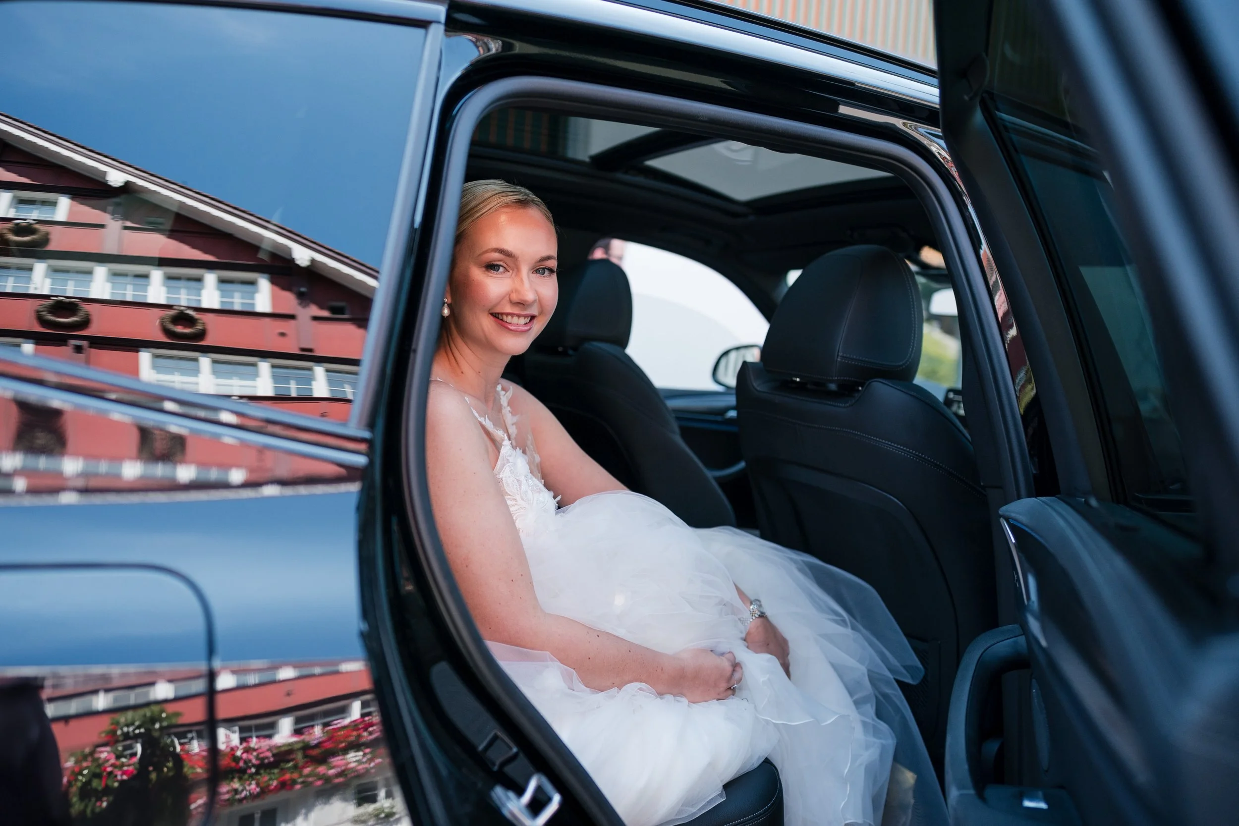 Eine Braut in einem weißen Brautkleid sitzt in einem schwarzen Auto, lächelt und schaut aus dem offenen Autofenster.
