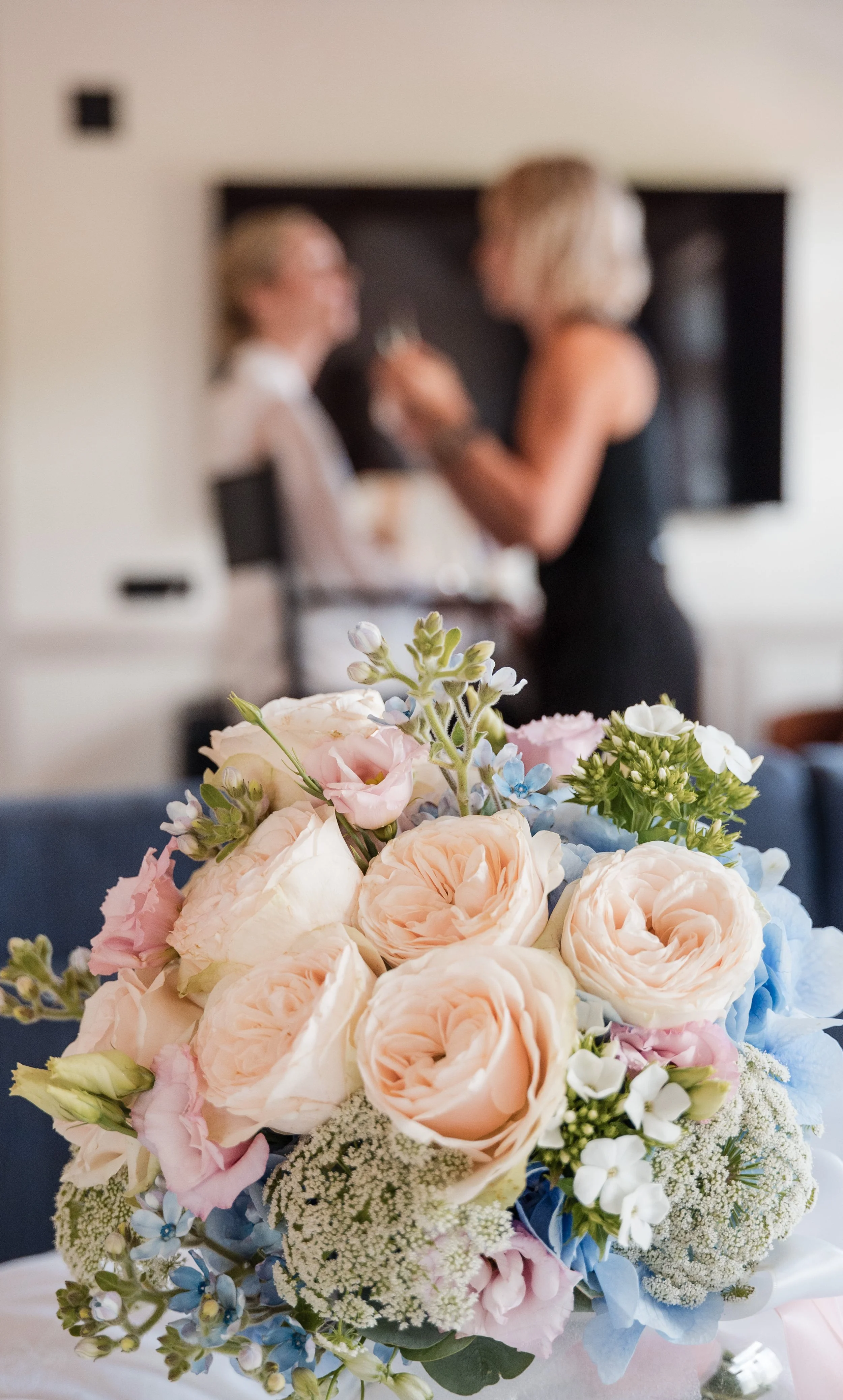 Bunte Blumenstrauß mit Rosen, Hortensien und anderen Blüten im Vordergrund, unscharfer Hintergrund mit zwei Frauen, die sich unterhalten