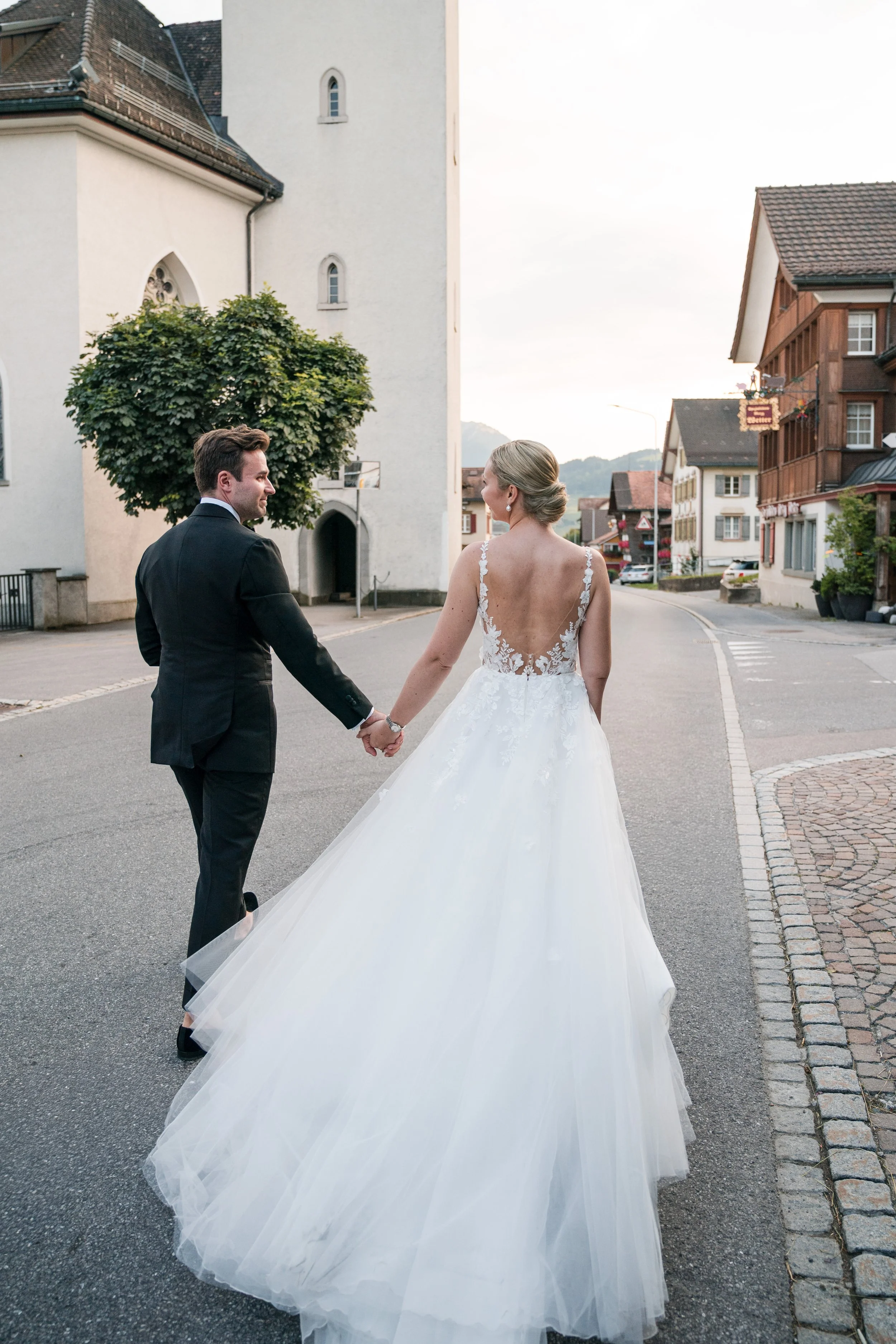 Braut und Bräutigam halten Händchen auf einer leeren Straße in einer kleinen Stadt, die Braut trägt ein weißes Hochzeitskleid mit offener Rückseite, der Bräutigam ein schwarzes Anzug, im Hintergrund alte Gebäude und eine Kirche, Abendstimmung.