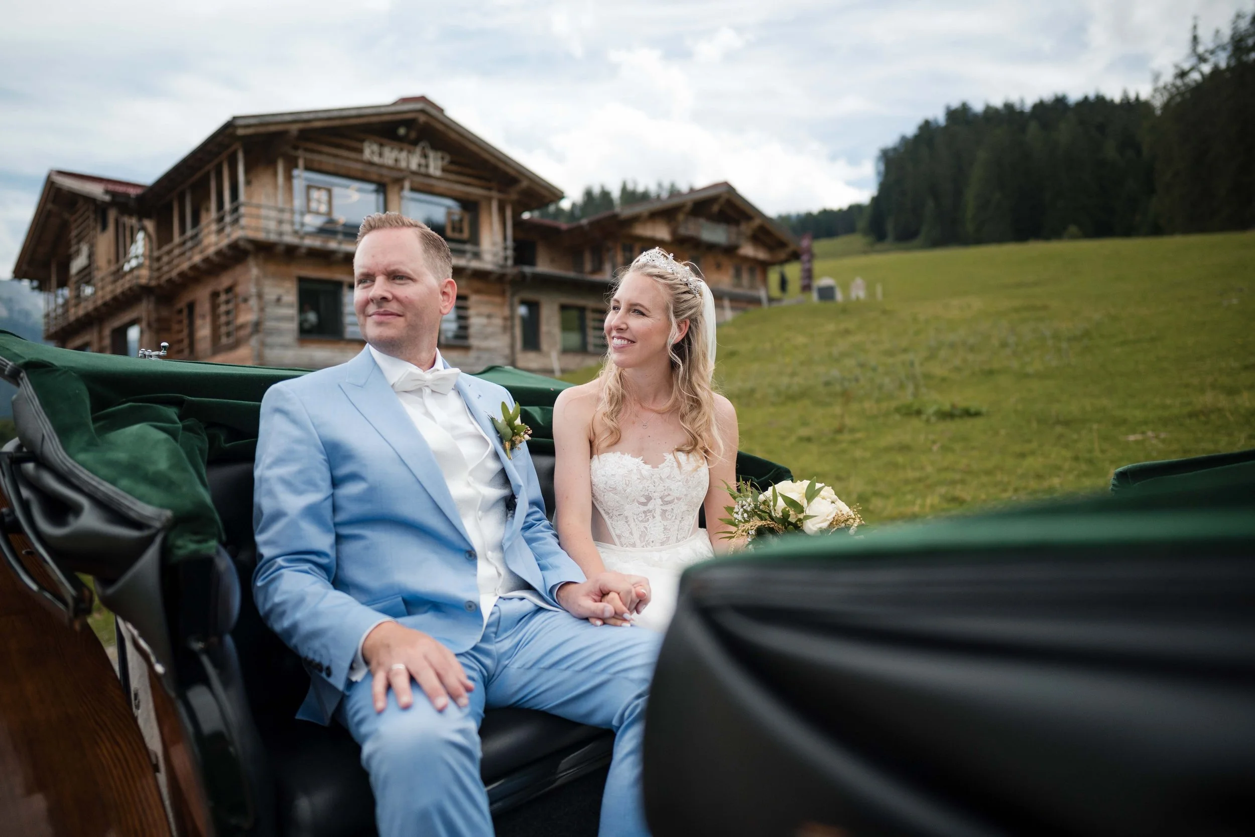 Ein frisch vermähltes Brautpaar sitzt in einer Kutsche vor einem holzgeschnitzten Haus in den Bergen, umgeben von grünen Wiesen, bei bewölktem Himmel.