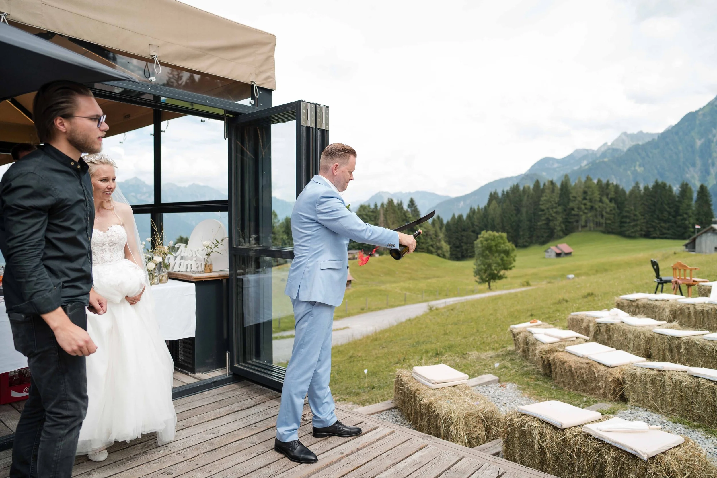 Ein Mann in einem blauen Anzug hält eine Flasche Wein und ein Glas, während er eine Rede bei einer Hochzeit im Freien vor einer Gruppe von Gästen hält. Ein Brautpaar steht im Hintergrund, umgeben von malerischer Landschaft mit Bergen und Wäldern.