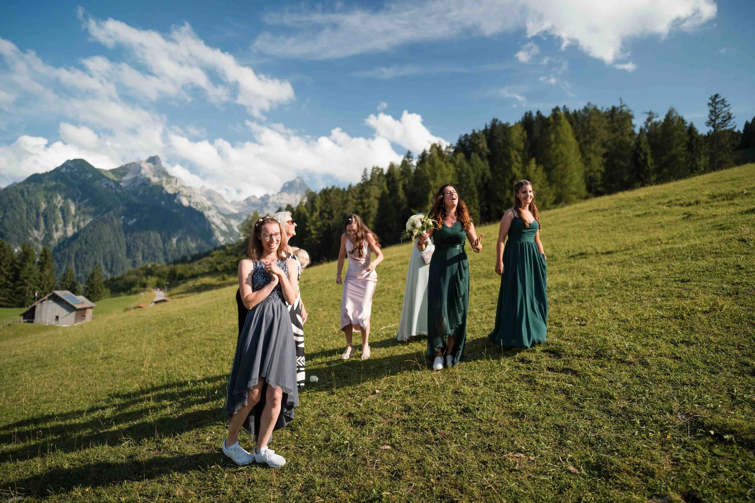 Mehrere Frauen in eleganten Kleidern spazieren auf einer grünen Wiese mit Bergen und Wald im Hintergrund, sonniger Himmel mit wenigen Wolken.