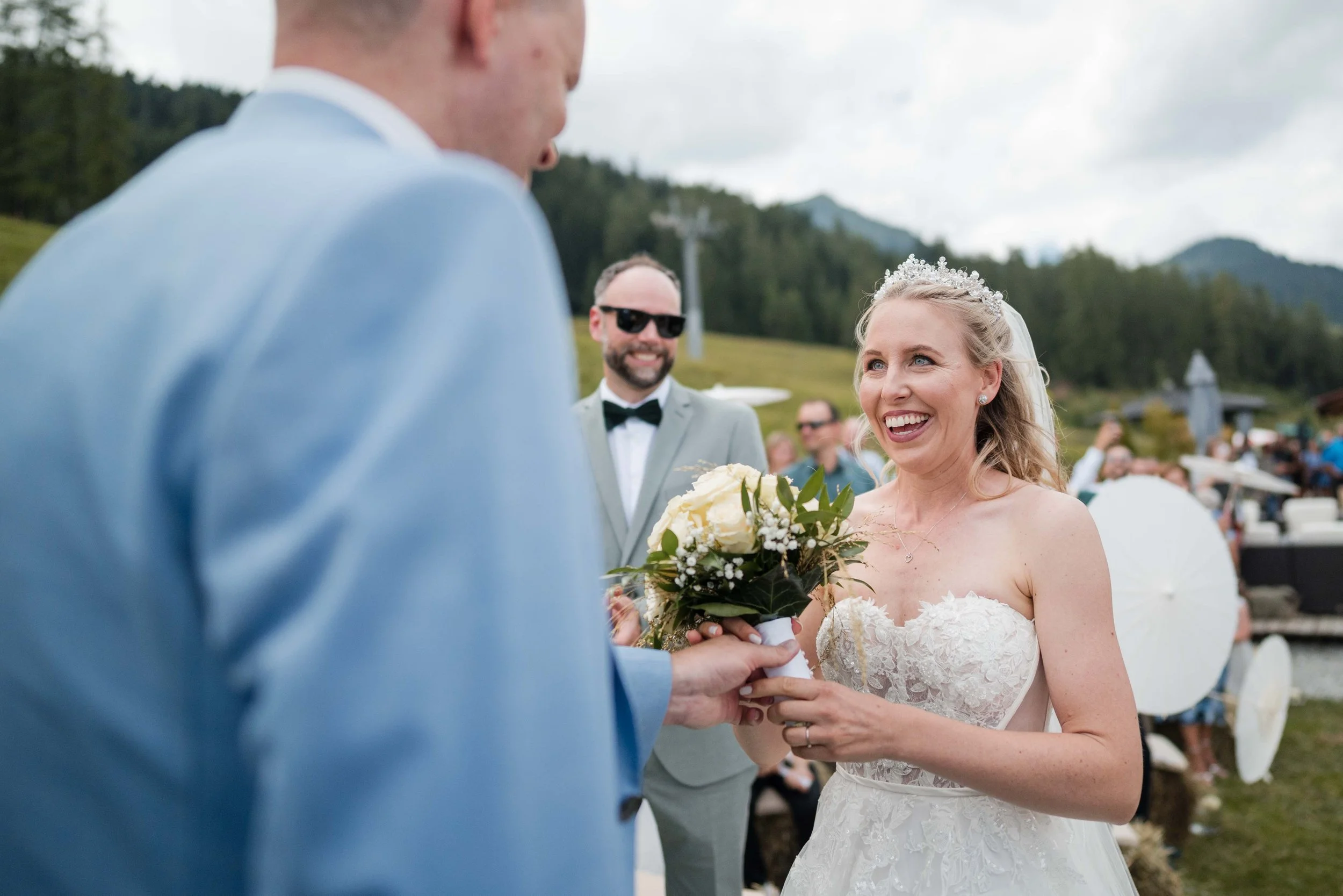 Braut in Hochzeitskleid lächelnd während der Trauung, hält Blumenstrauß, im Hintergrund Gäste und grüner Berg-Hintergrund.
