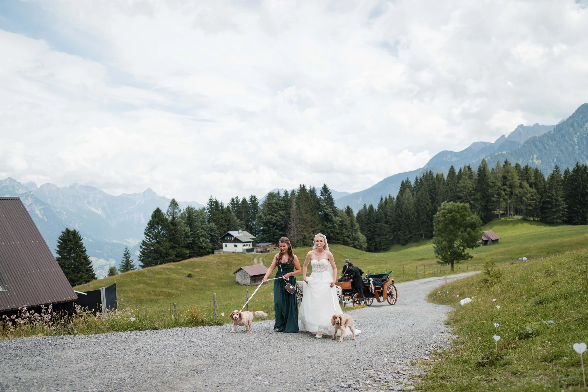 Zwei Frauen in Brautkleidung gehen auf einer Landstraße, begleitet von zwei Hunden, während im Hintergrund eine Kutsche mit zwei Personen vorbeifährt und eine malerische Berglandschaft zu sehen ist.