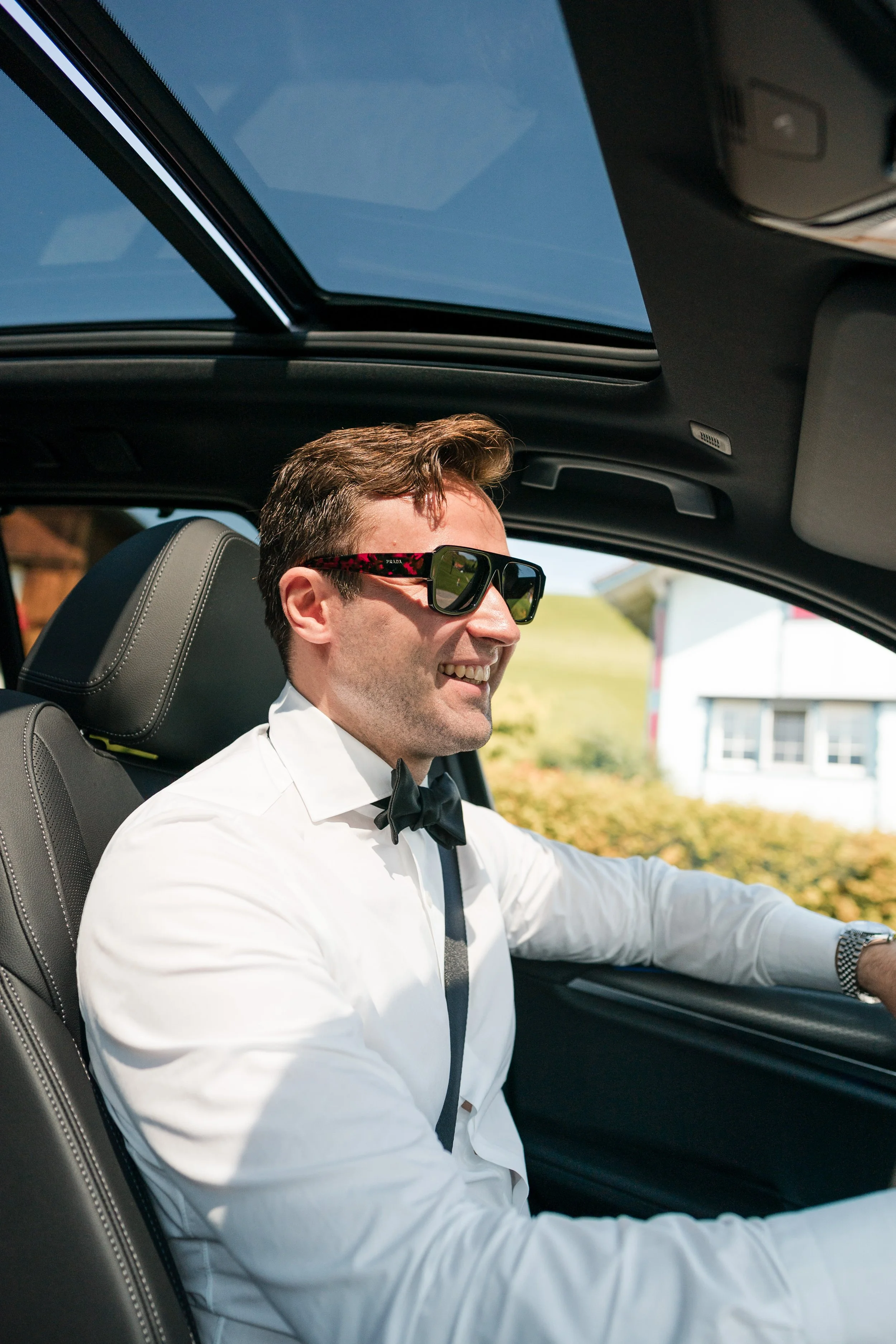 Ein Mann in einem weißen Hemd mit Fliege sitzt in einem Auto, trägt Sonnenbrille und lächelt, während er fährt