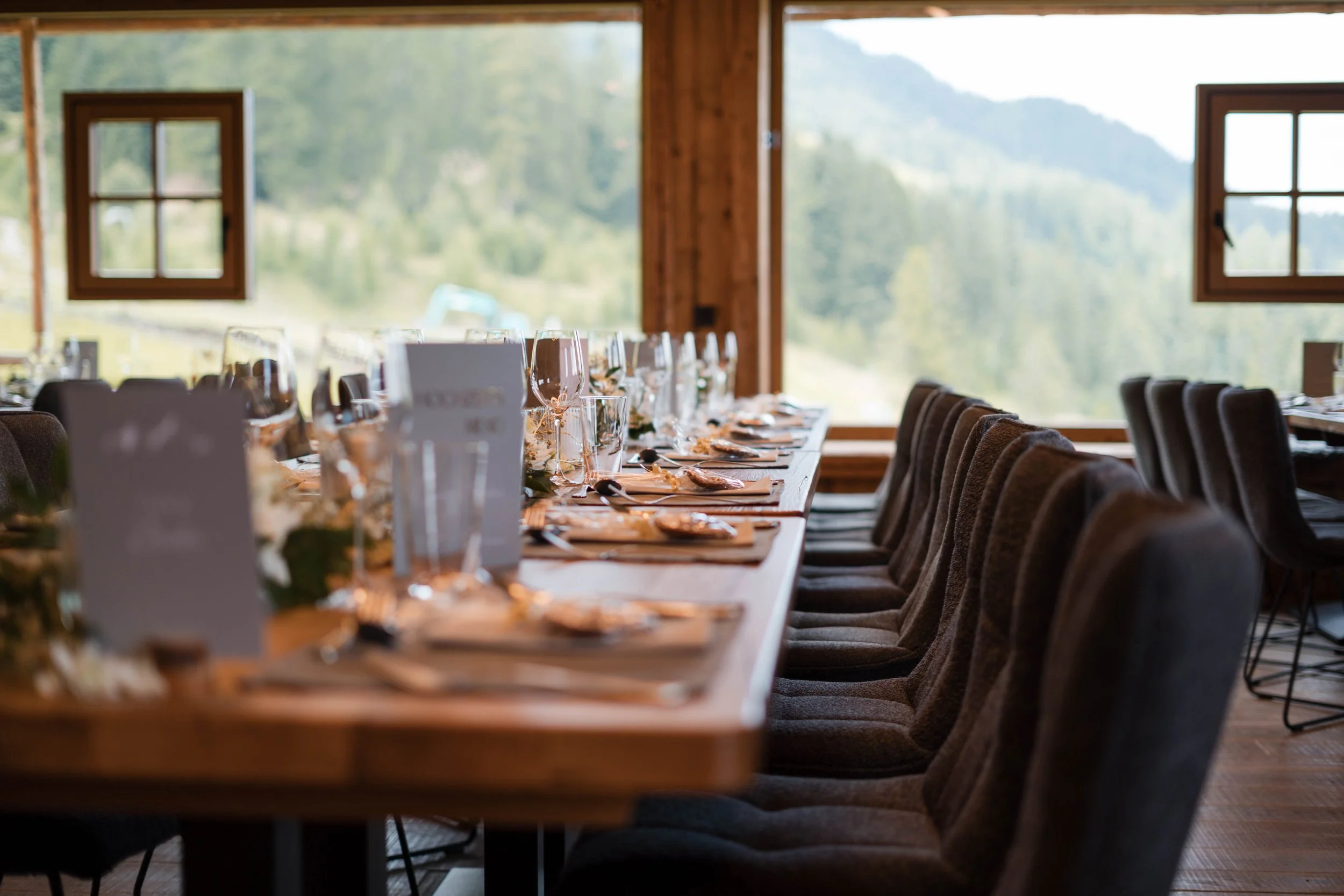 Ein festlich gedeckter Esstisch in einer rustikalen Holzhütte mit Blick auf eine grüne Berglandschaft, inklusive Gläser, Teller und Menükarten.