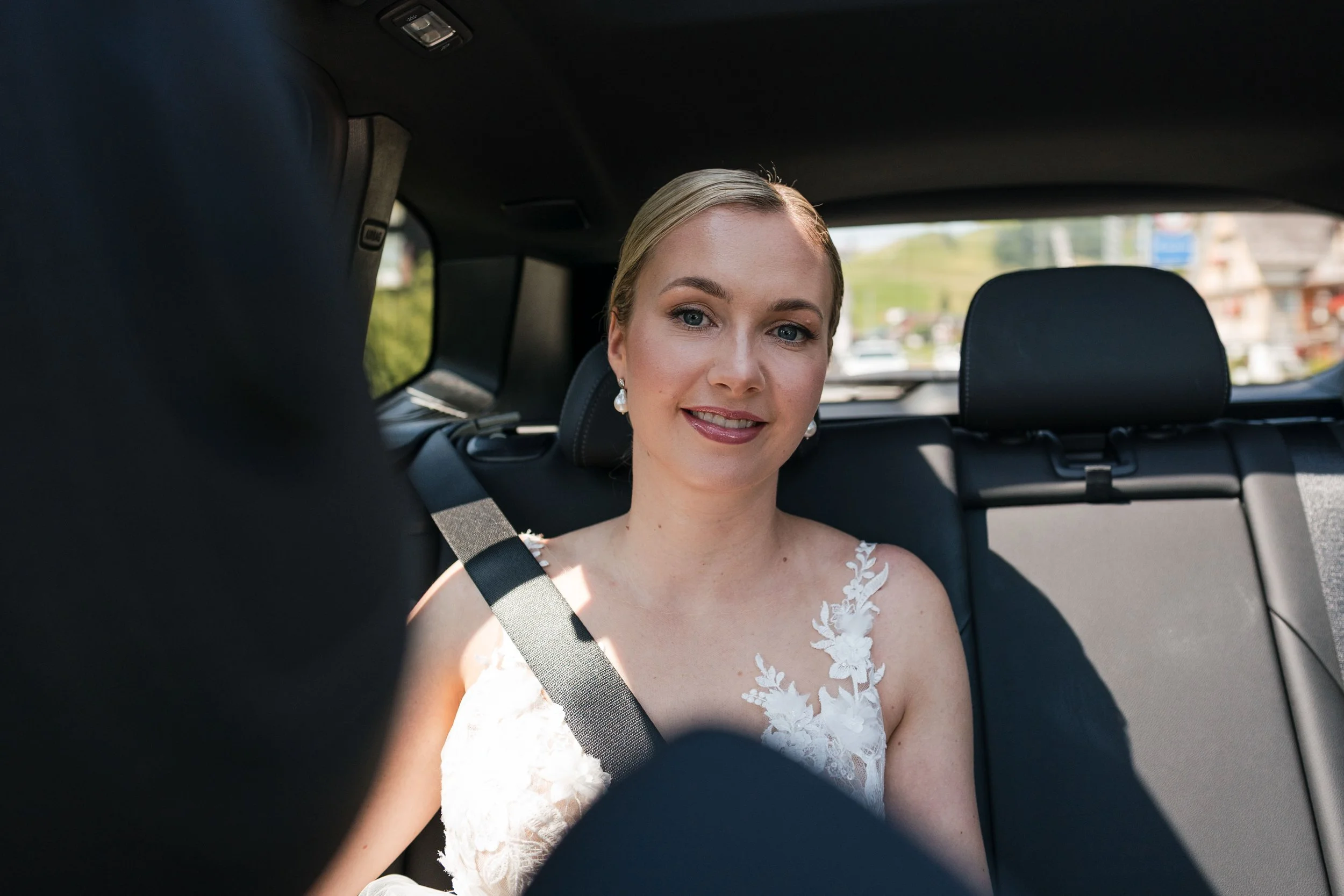 Eine junge Frau im weißen Hochzeitskleid sitzt auf dem Rücksitz eines Autos, mit Hellblonden Haaren, die zu einem Knoten gebunden sind, und trägt Perlenohrringe. Sie lächelt freundlich in die Kamera. Im Hintergrund ist die Straße und grüne Landschaft