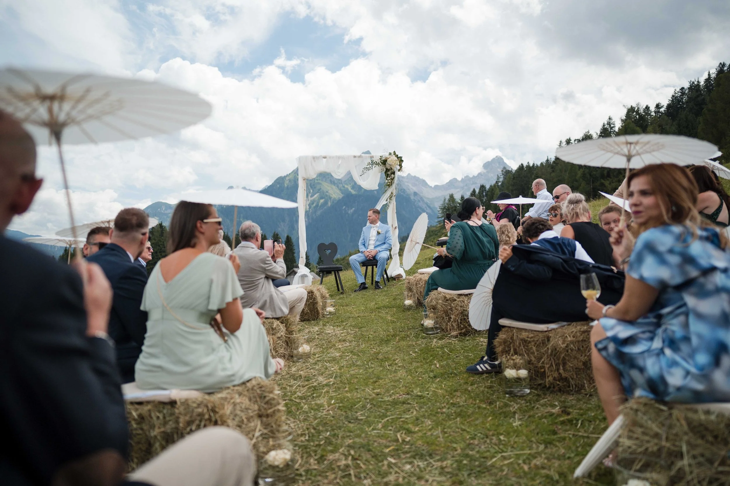 Hochzeit im Freien in den Bergen, Gäste sitzen auf Strohballen mit Sonnenparaphern, im Hintergrund ein Bergpanorama, Brautpaar vor einem weißen Bogen, einige Gäste halten Fotos.