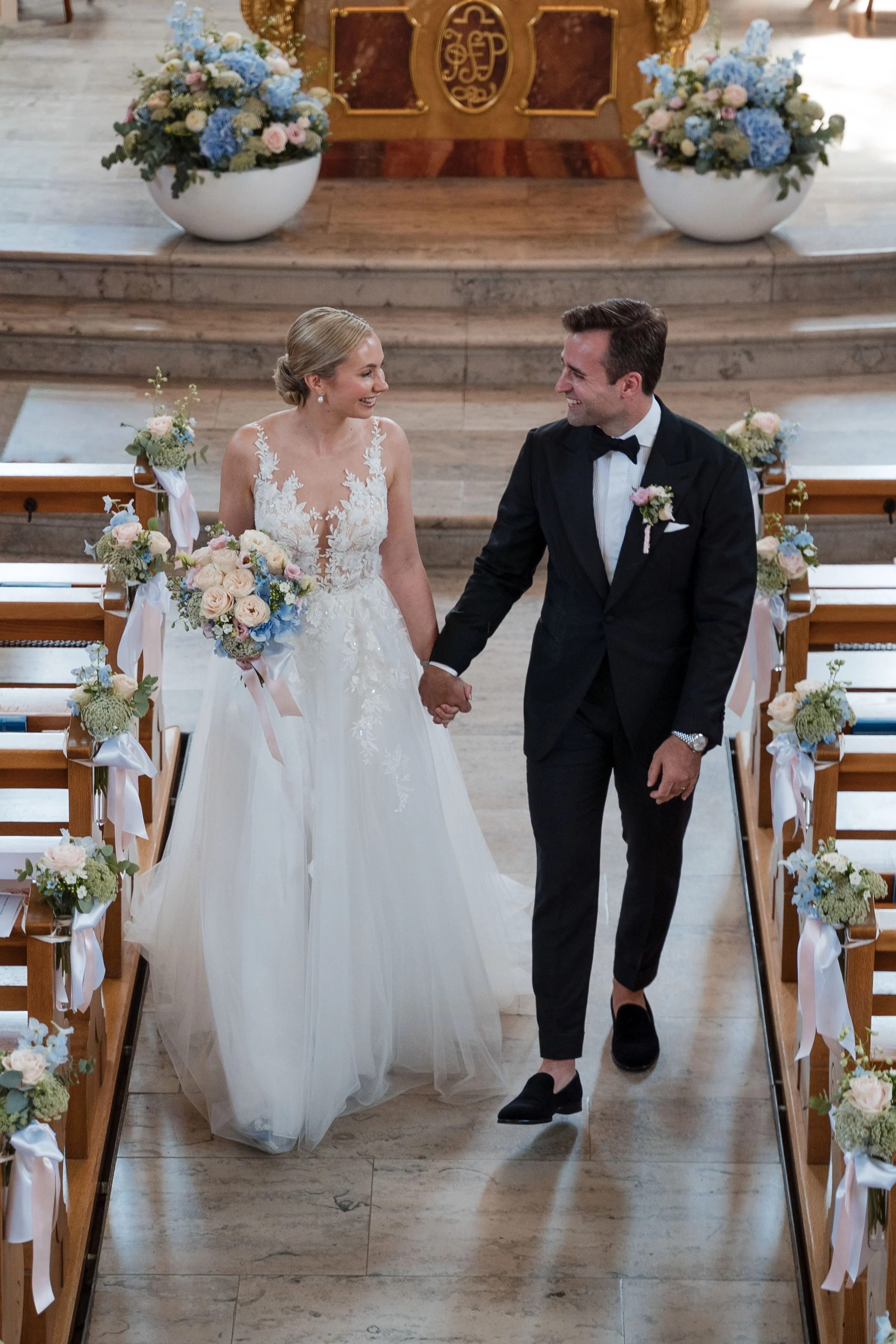 Ein frisch verheiratetes Paar hält Händchen in einer Kirche, umgeben von Blumen, während sie auf den Altar zulaufen.