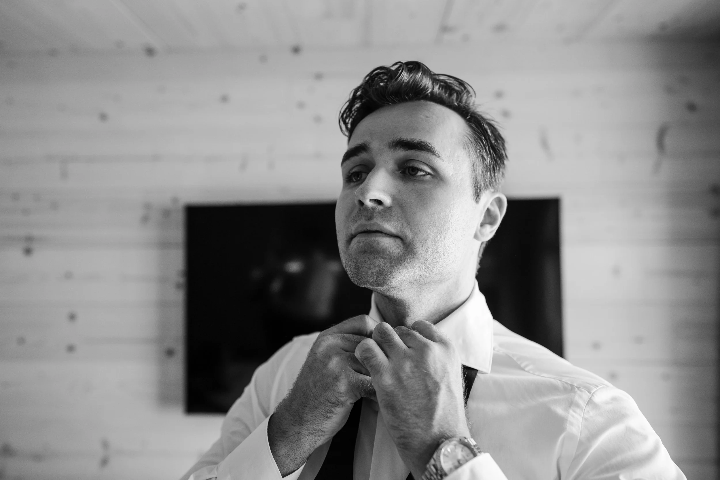 Schwarz-Weiß-Foto eines jungen Mannes, der an einem Hemd und Krawatte schraubt, ohne Krawatte zu tragen, beim Vorbereiten auf ein formelles Ereignis.