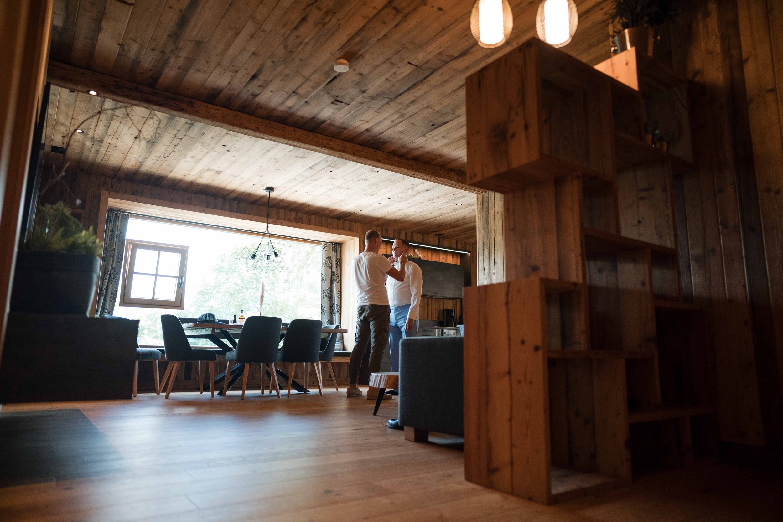 Zwei Männer im Gespräch in einem holzverkleideten Wohnzimmer, mit einem großen Esstisch und Stühlen vor einem Fenster.