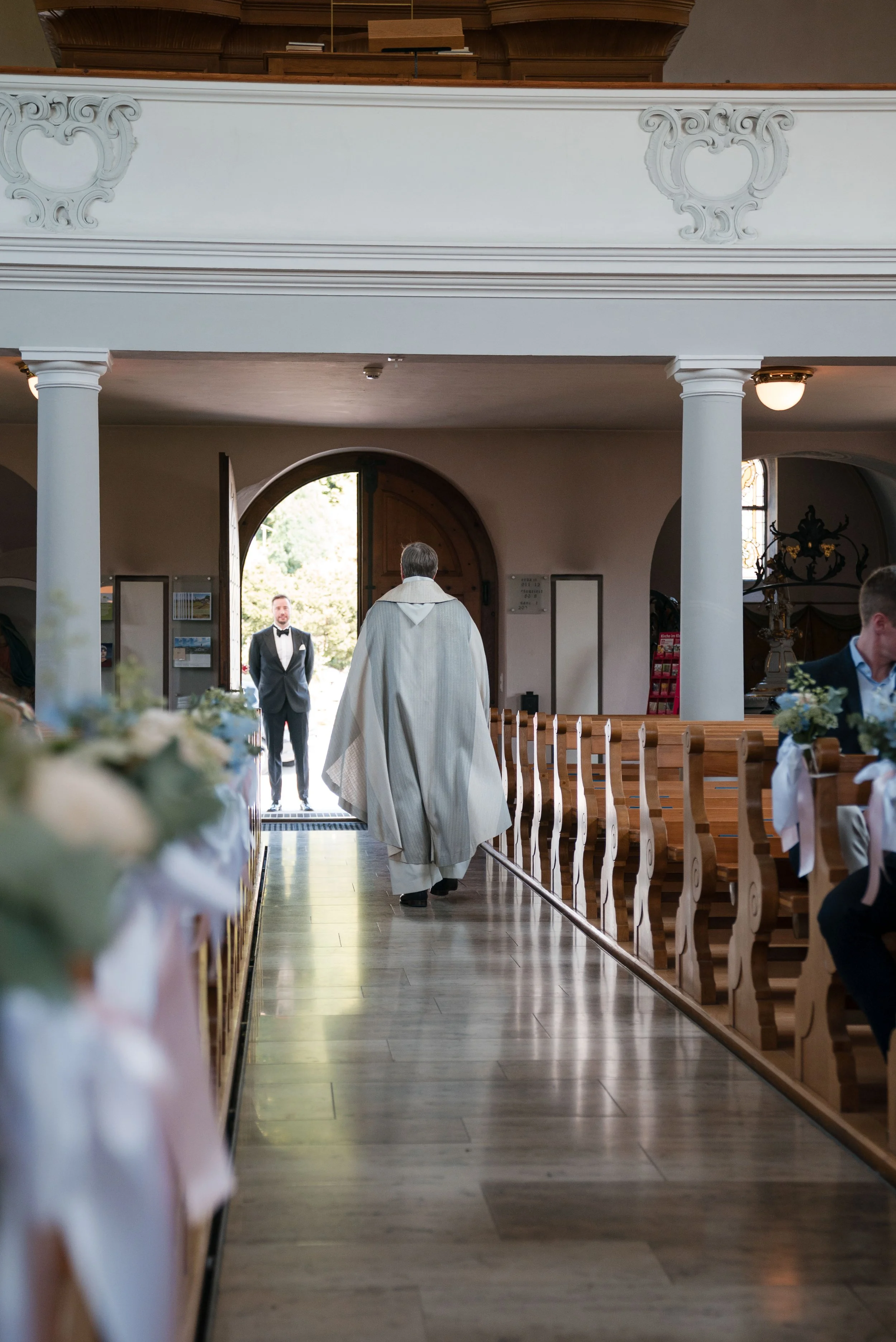 Ein Priester in weißer albenartiger Robe betreten die Kirche, während ein Bräutigam in smokingartiger Kleidung im Hintergrund vor der Tür steht. Die Kirche ist elegant mit Holzbänken und dekorativen Elementen ausgestattet.