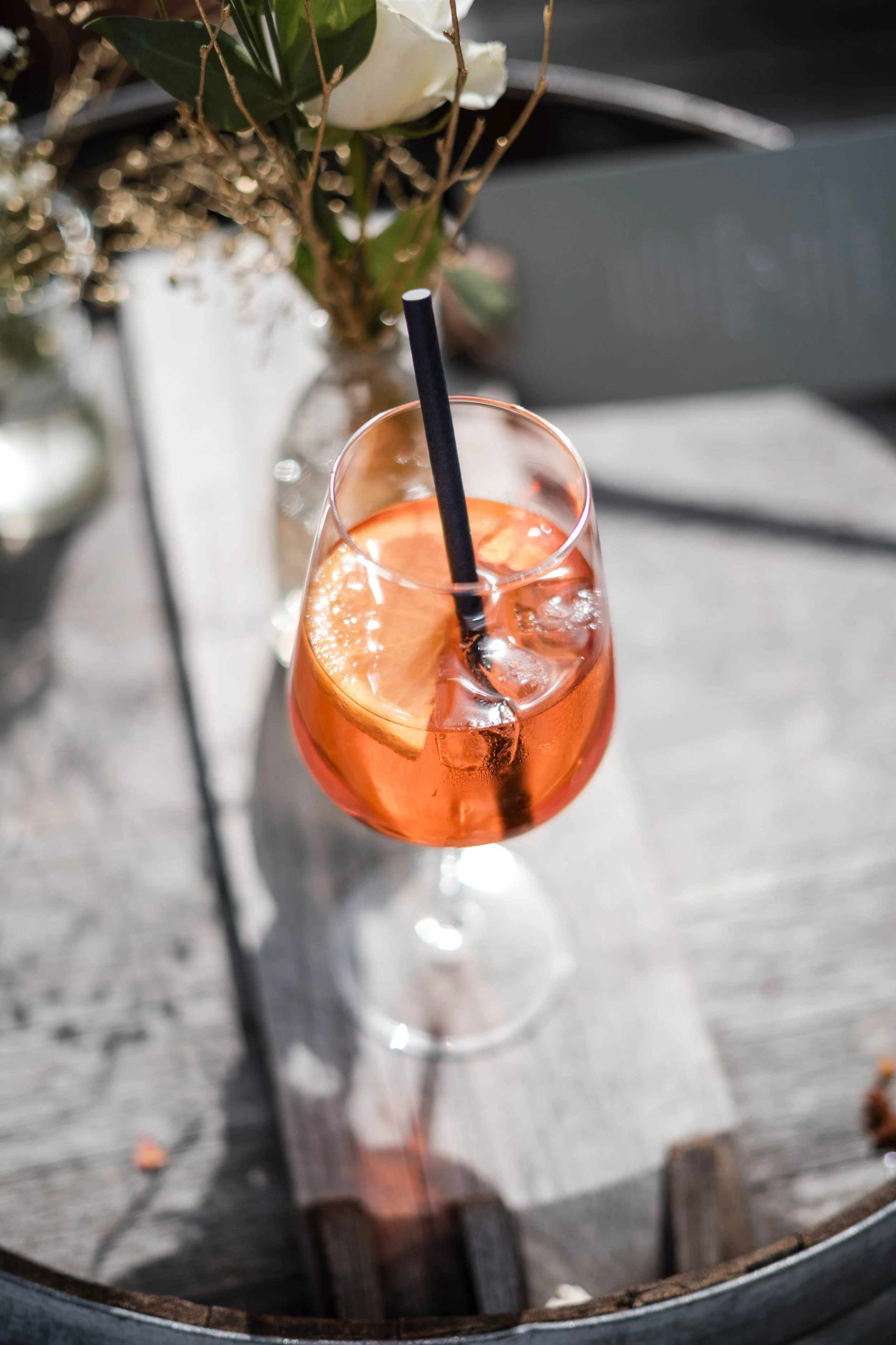 Ein Glas mit rotem Getränk, Eiswürfeln und einer Zitronenscheibe, mit einem schwarzen Strohhalm, auf einem Holztisch mit Blumen im Hintergrund.