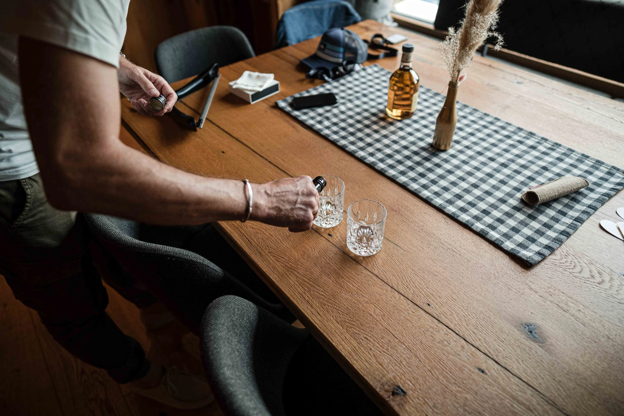 Eine Person stellt zwei Gläser auf einen Holztisch, auf dem auch eine Flasche, eine Vase mit getrockneten Pflanzen, eine Jacke, eine Cap und andere Gegenstände liegen.