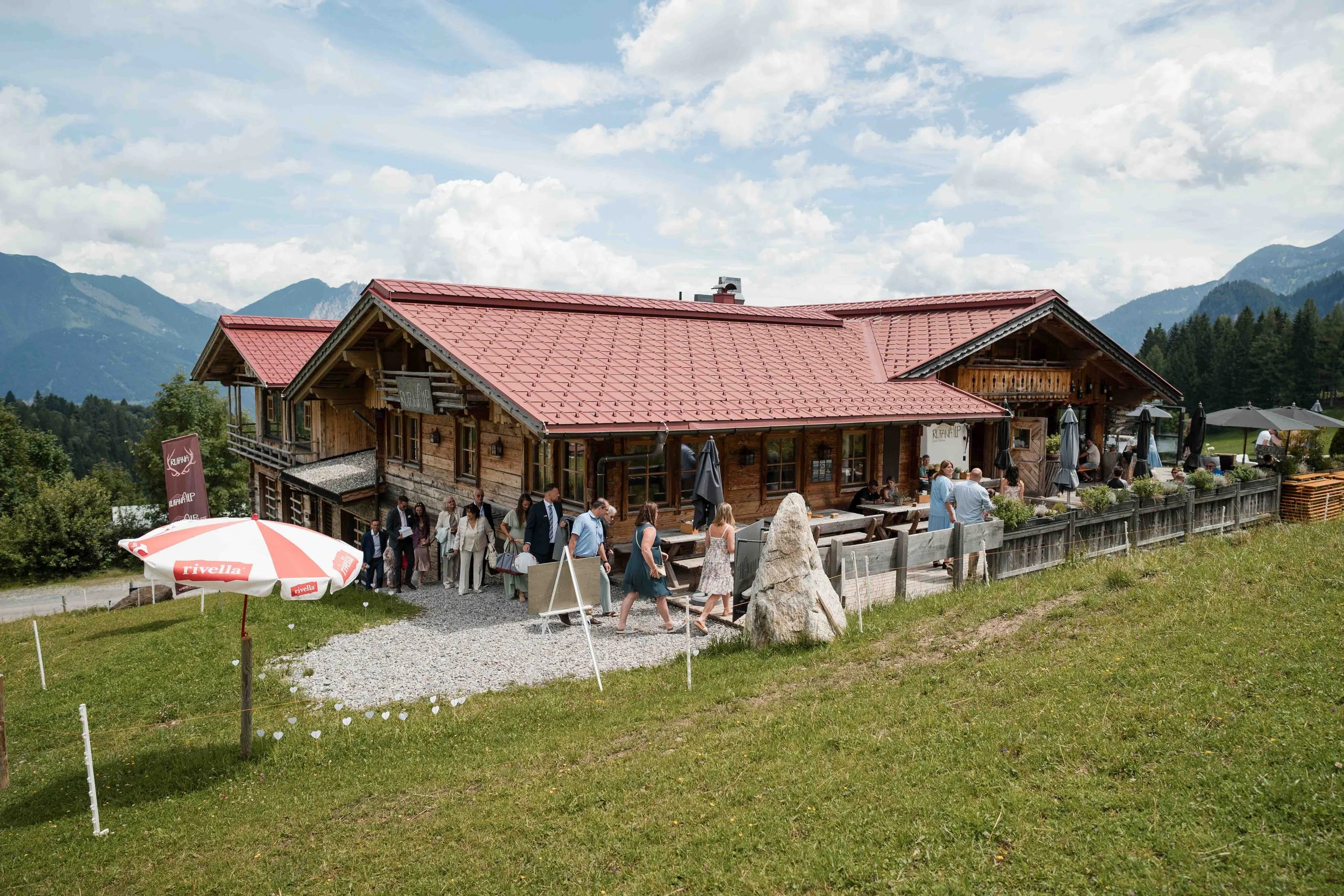 Ein Holzrestaurant mit roten Dächern in einer bergigen Landschaft. Im Freien sitzen Menschen und gehen durch das Gebiet. Es gibt einen weißen Sonnenschirm mit dem Namen »rivella« und eine große Felsbrocken auf der Terrasse.