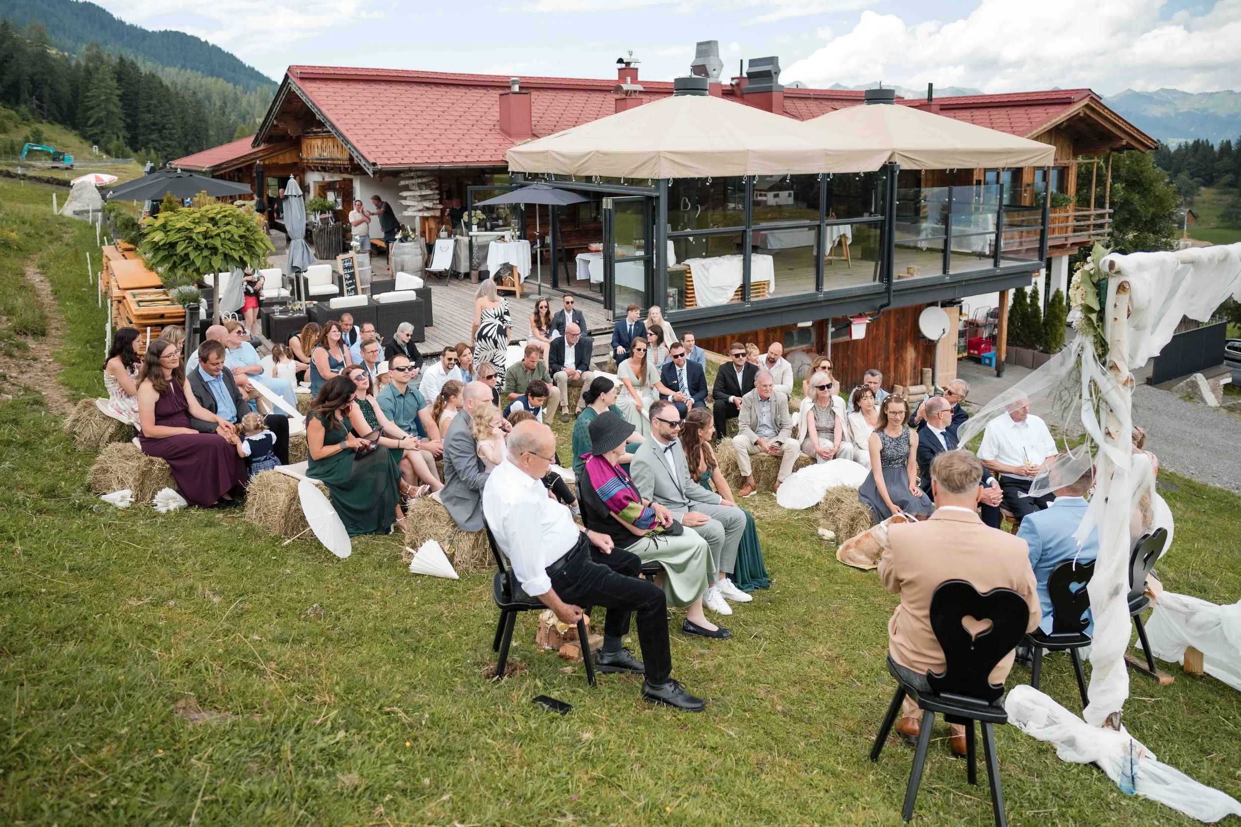 Eine outdoor Hochzeitszeremonie mit vielen Gästen, die auf Strohballen vor einem Chalet in den Bergen sitzen.