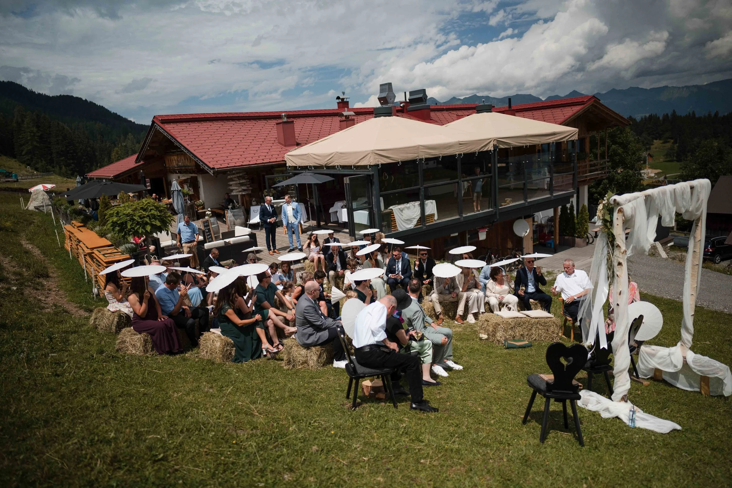 Outdoor Hochzeitszeremonie auf einer Wiese vor einem Chalet mit Bergen im Hintergrund, Gäste mit weißen Sonnenschirmen sitzen auf Strohballen, ein Redner bei der Zeremonie, dekorierter Hochzeitsbogen aus Stoff und Holz.