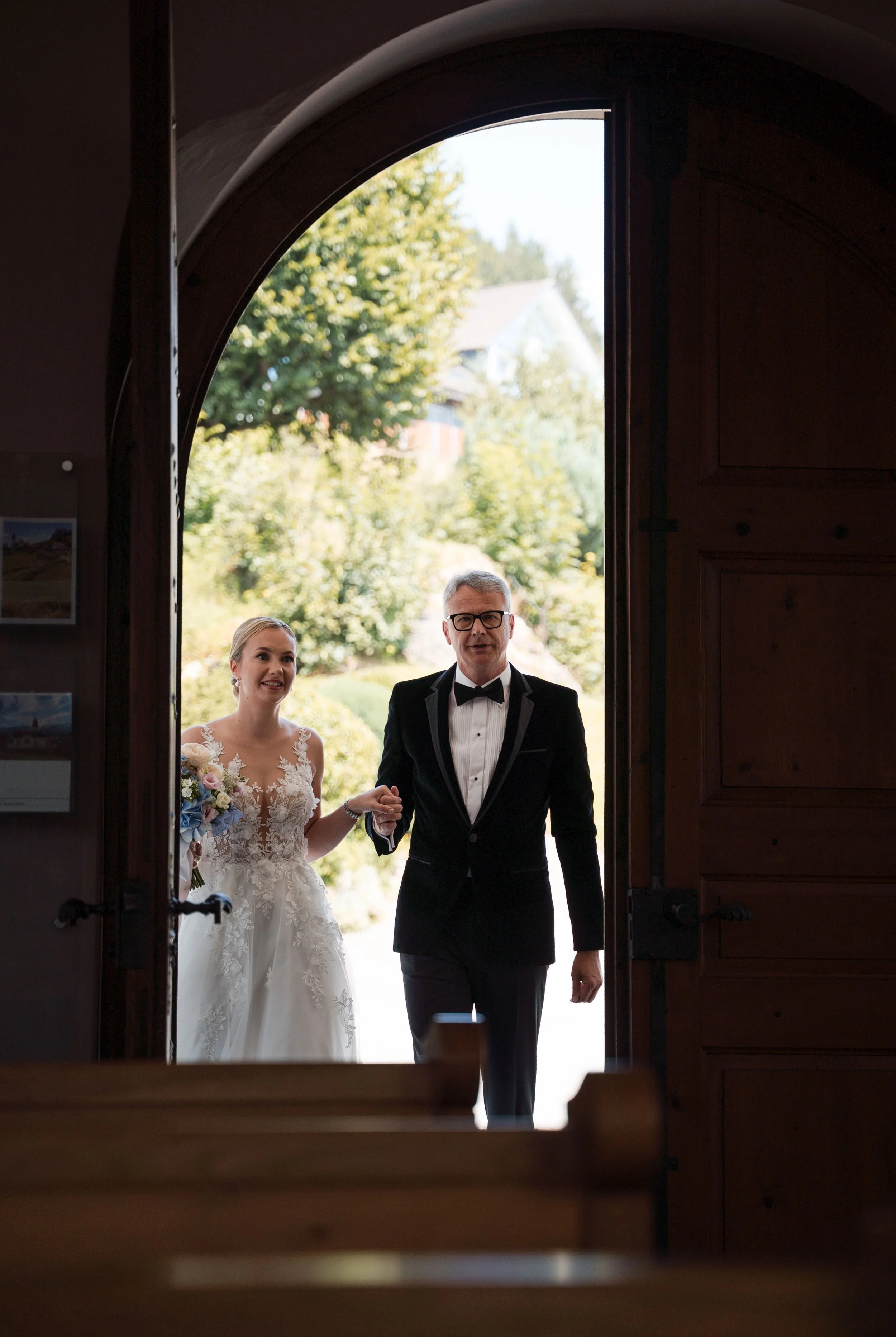 Braut und Vater betreten Kirche beim Hochzeitsgottesdienst, draußen grüne Bäume und ein Haus sind sichtbar.