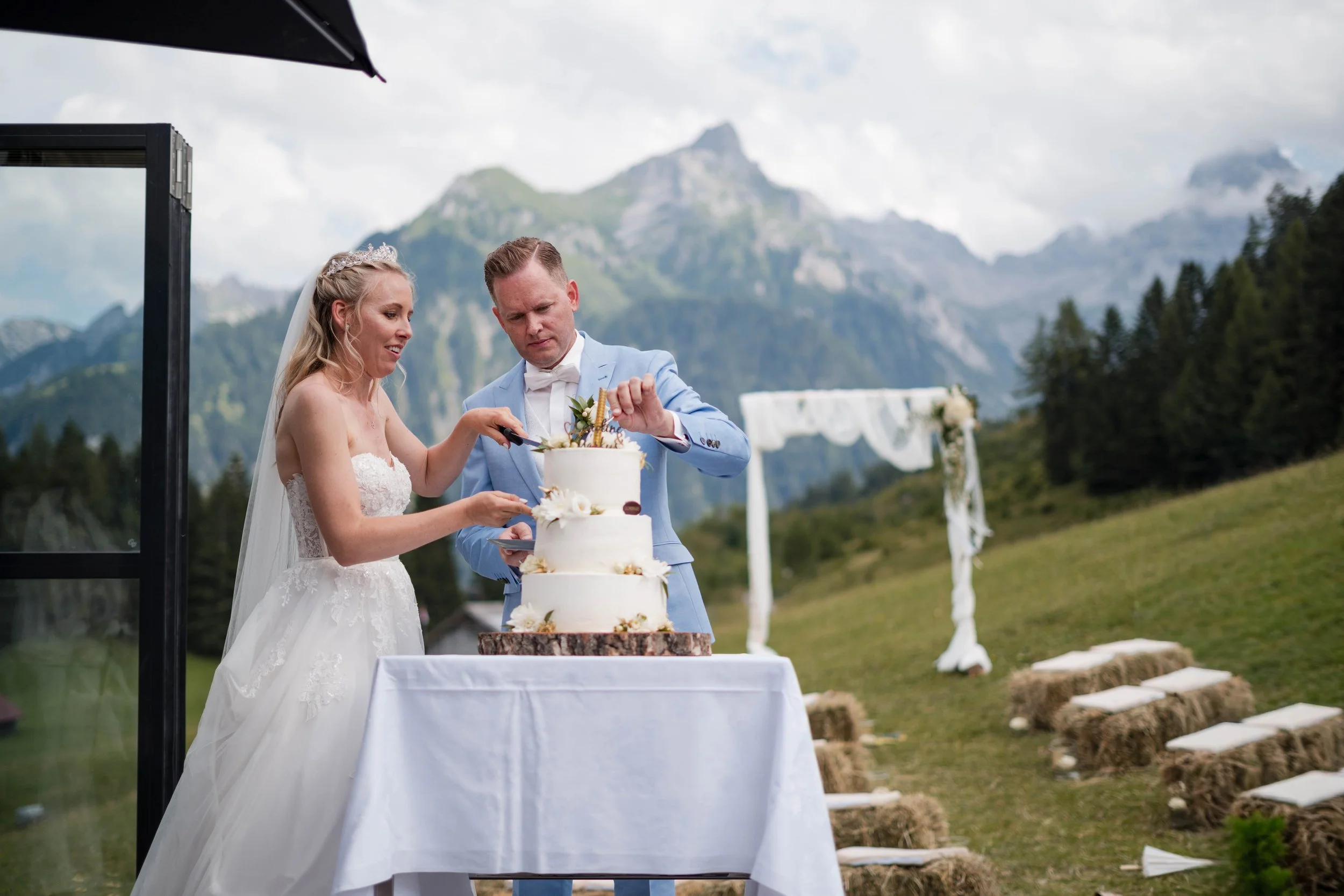 Ein Brautpaar schneidet eine Hochzeitstorte im Freien mit Bergen im Hintergrund.