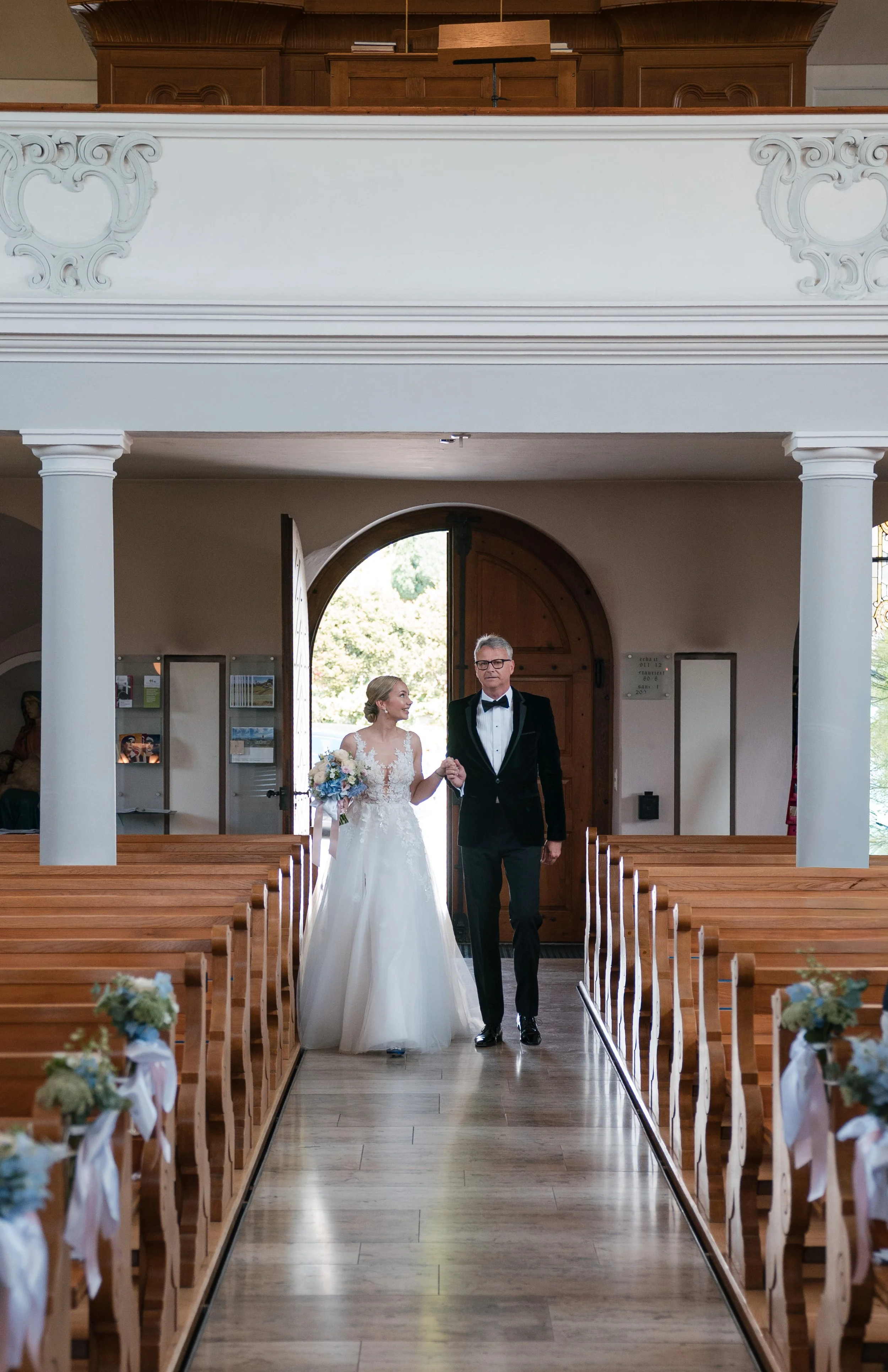 Braut und Vater betreten eine Kirche während einer Hochzeit, dekorierte Holzbänke, offene Tür mit Blick nach draußen.