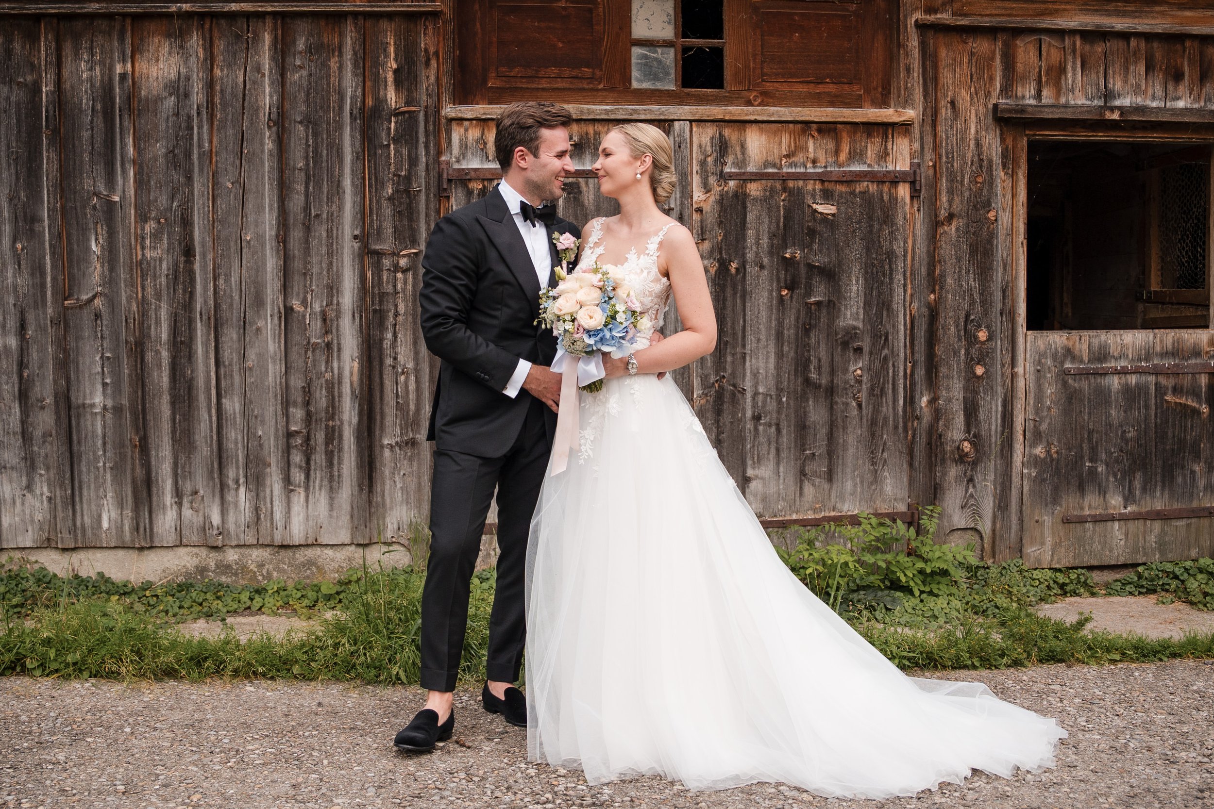 Braut und Bräutigam stehen sich vor einer alten Holzscheune, lachen sich an, die Braut hält einen Blumenstrauß und trägt ein weißes Brautkleid, der Bräutigam trägt einen schwarzen Anzug.