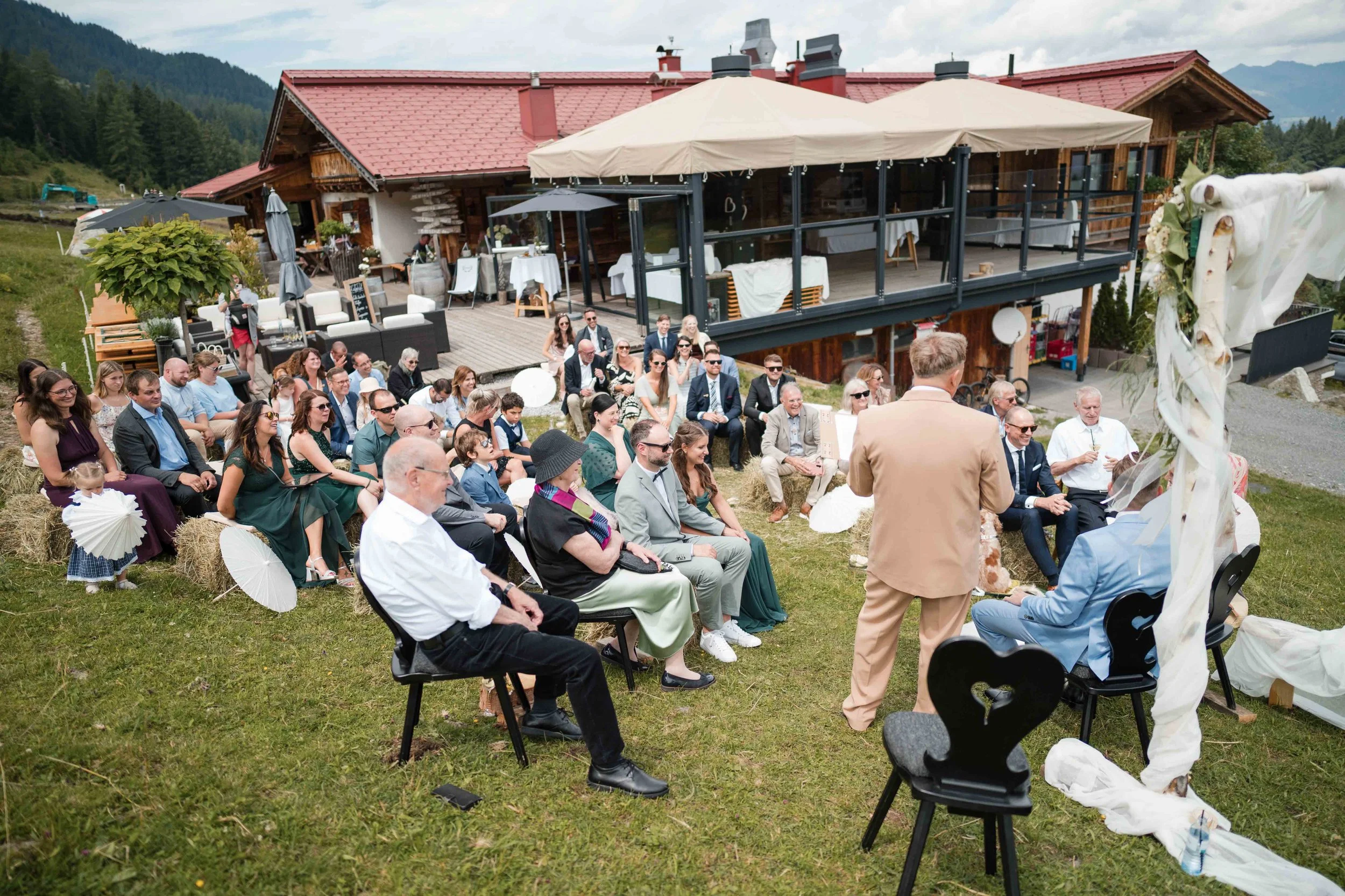 Menschen nehmen an einer outdoor Hochzeit auf einer Wiese vor einem rustikalen Holzhaus teil, mit Strohballen, Sonnenschirmen und Dekorationen, bei schönem Wetter.