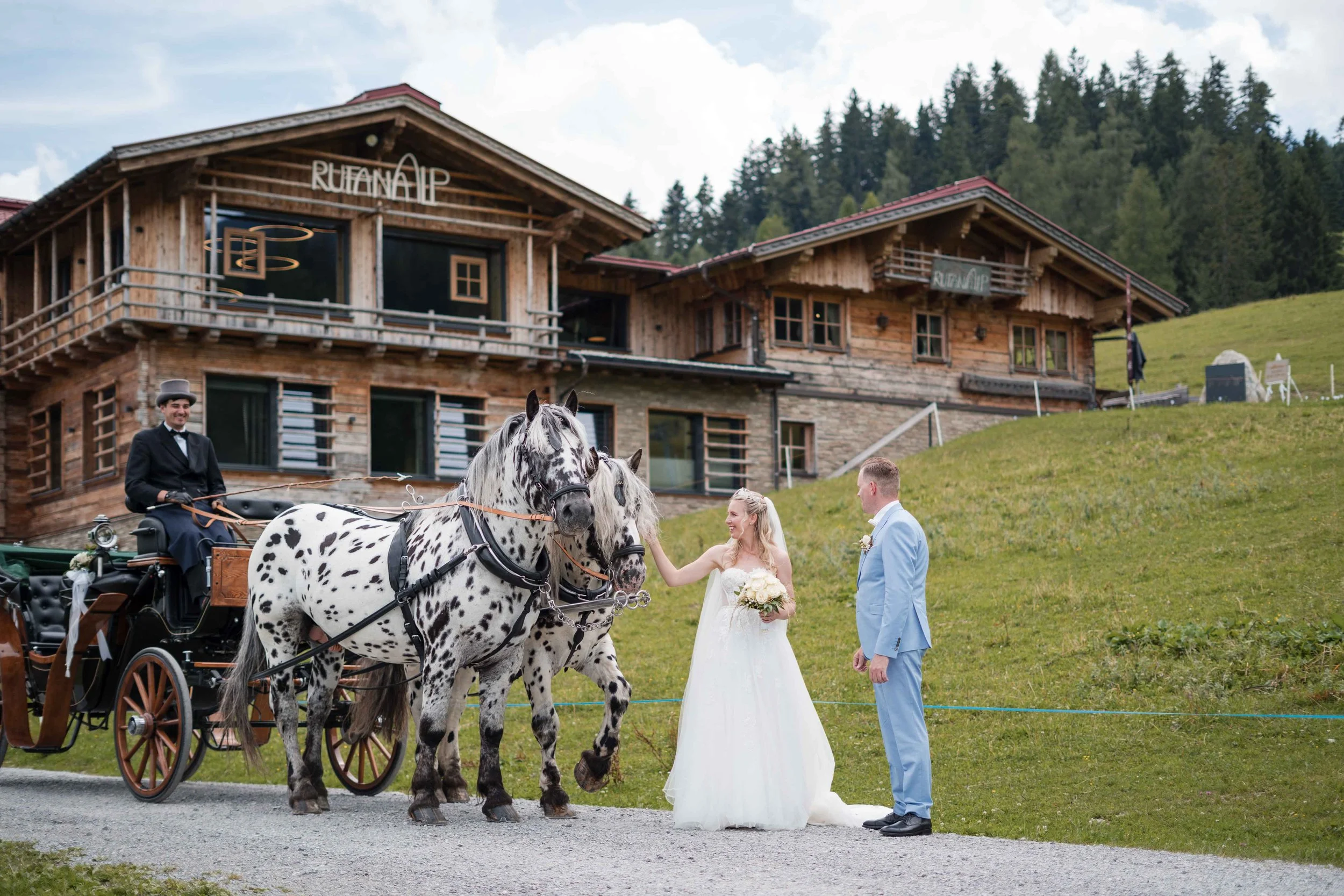 Ein Brautpaar mit einem Kutscher auf einer Pferdekutsche vor einem Holzhaus auf einer grünen Wiese, beim Heiratszeremonie, in den Bergen, mit bewölktem Himmel.