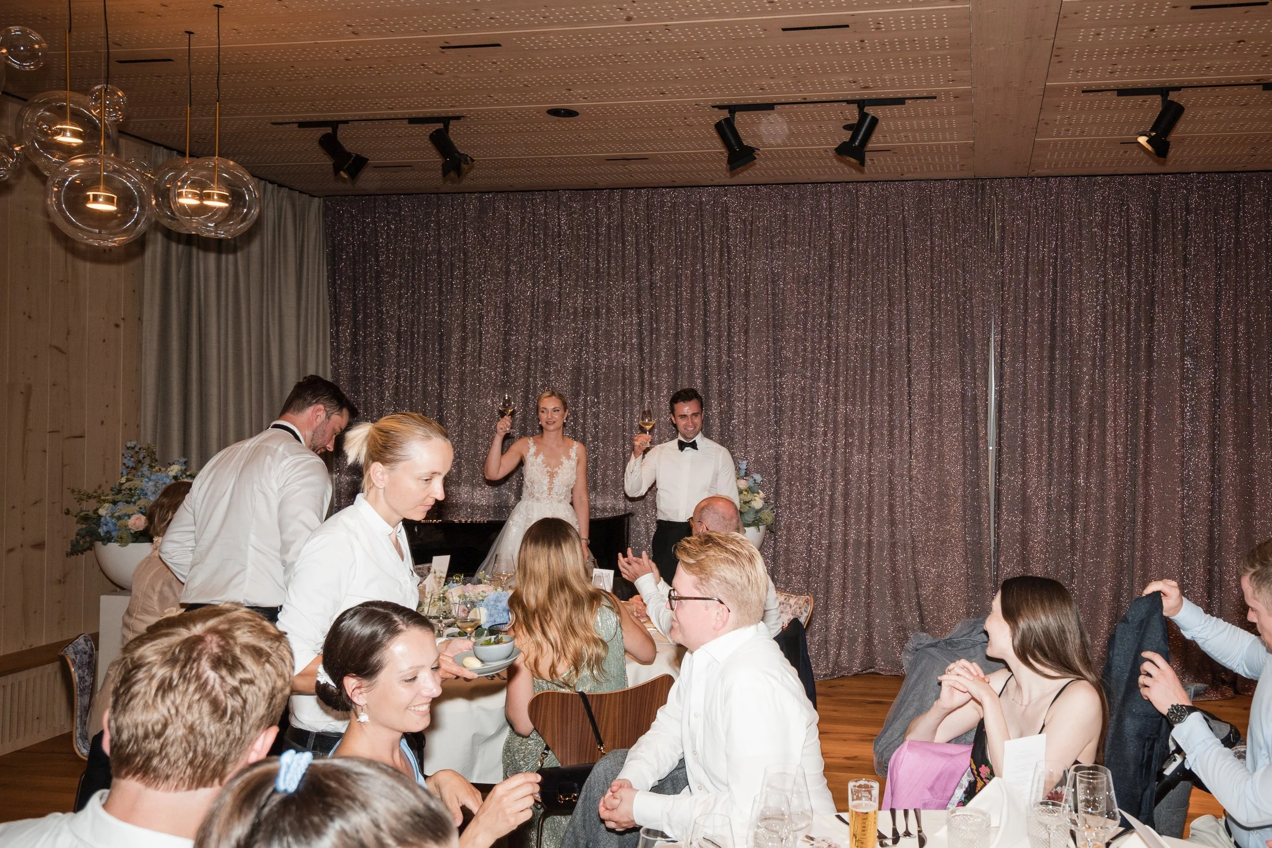 Hochzeitspaar bei der Feier, Gäste sitzen am Tisch, Servierer bedienen, festliche Dekoration, Bühne mit Glitter-Hintergrund.