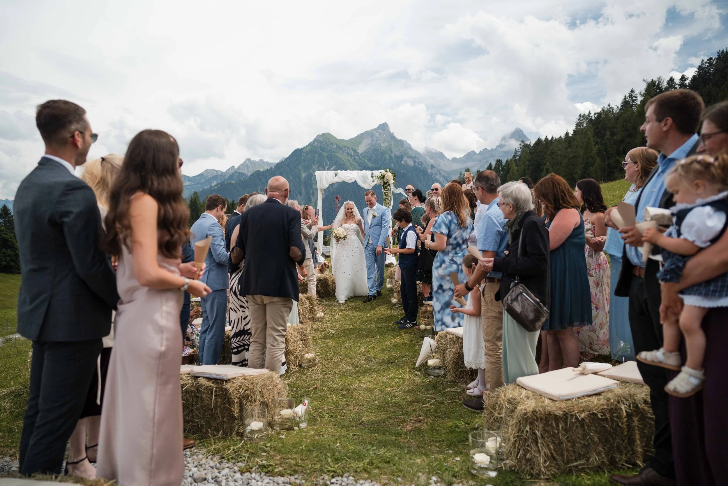 Eine Hochzeitsfeier im Freien inmitten von Bergen mit einer Hochzeitsinstalation, bei der ein Brautpaar vor Gästen eine Zeremonie durchführt.
